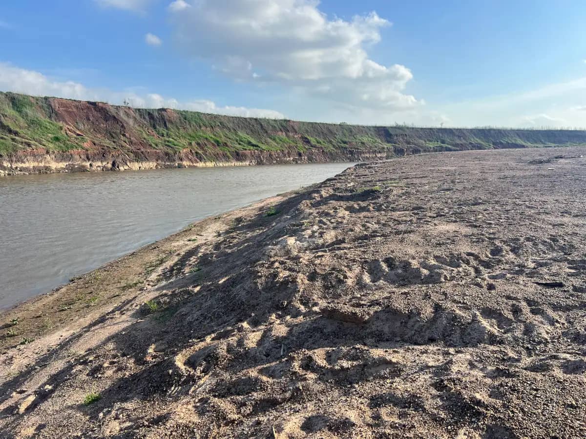 A perspective shot looking down a steep, rough sand-and-mud riverbank running parallel to the water toward a distant eroded cliff line.
