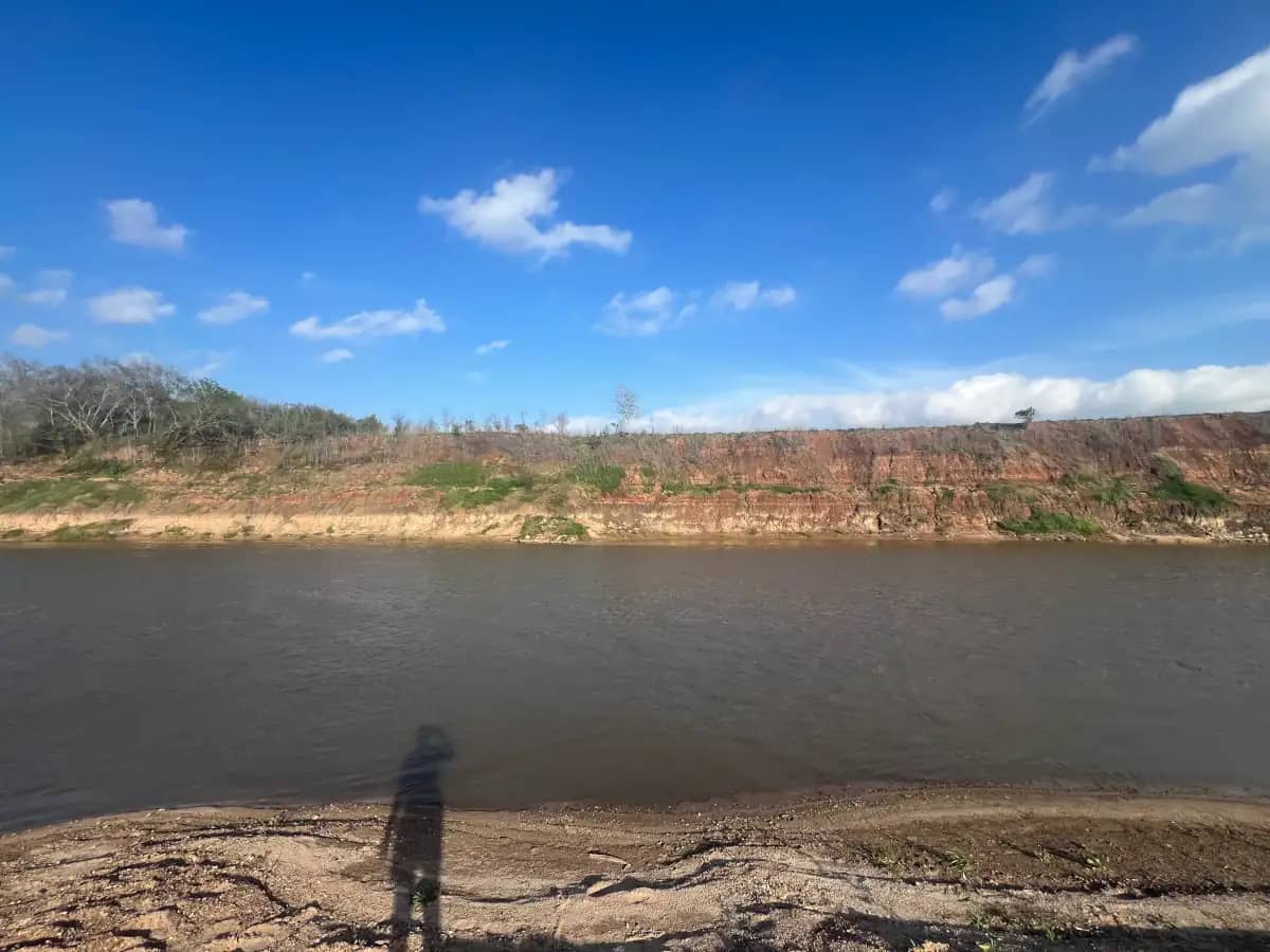 A direct shot looking straight across a wide brown river toward a steep dirt cliff face, with the long shadow of the photographer cast on the muddy bank.