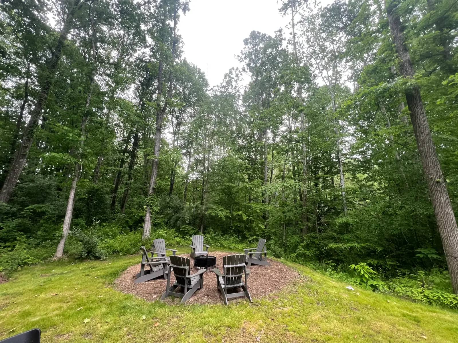 A circular mulch clearing tucked into a dense, green hardwood forest featuring a central black steel fire ring surrounded by six gray Adirondack chairs.