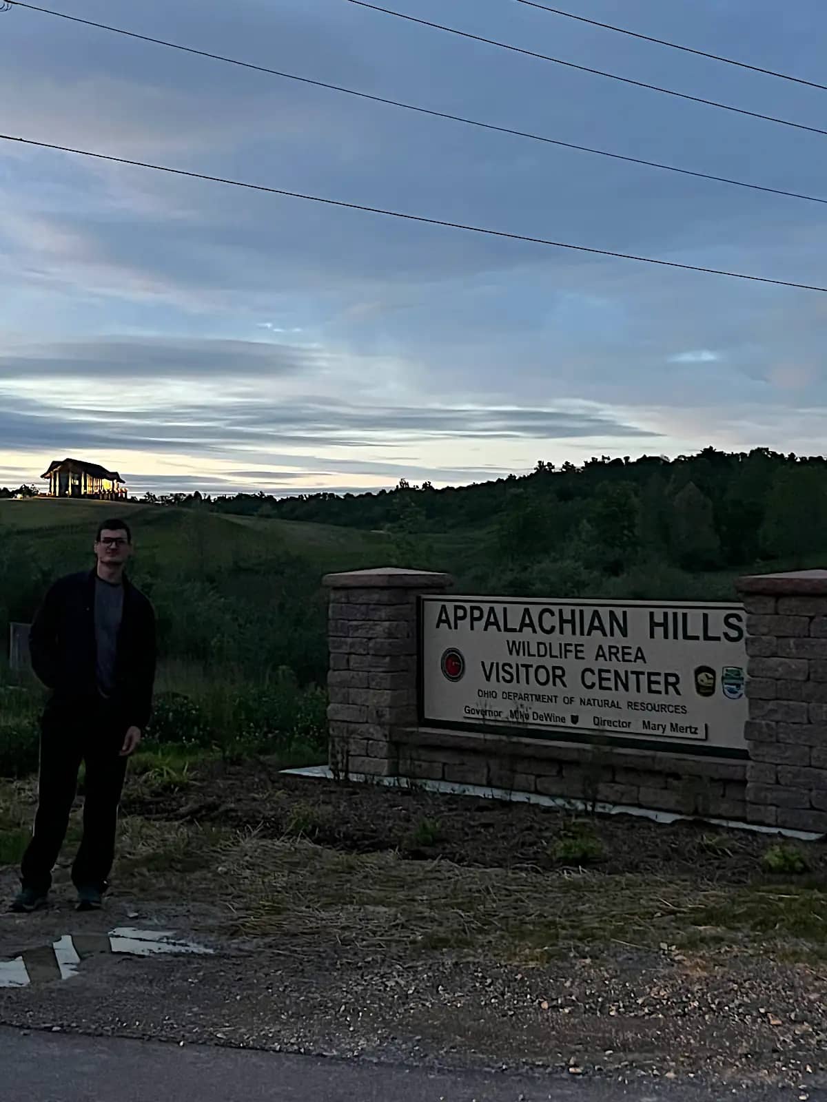 A man standing next to the stone-pillared entrance sign for the Appalachian Hills Wildlife Area Visitor Center, with the main building lit up on a distant hill under an evening sky.