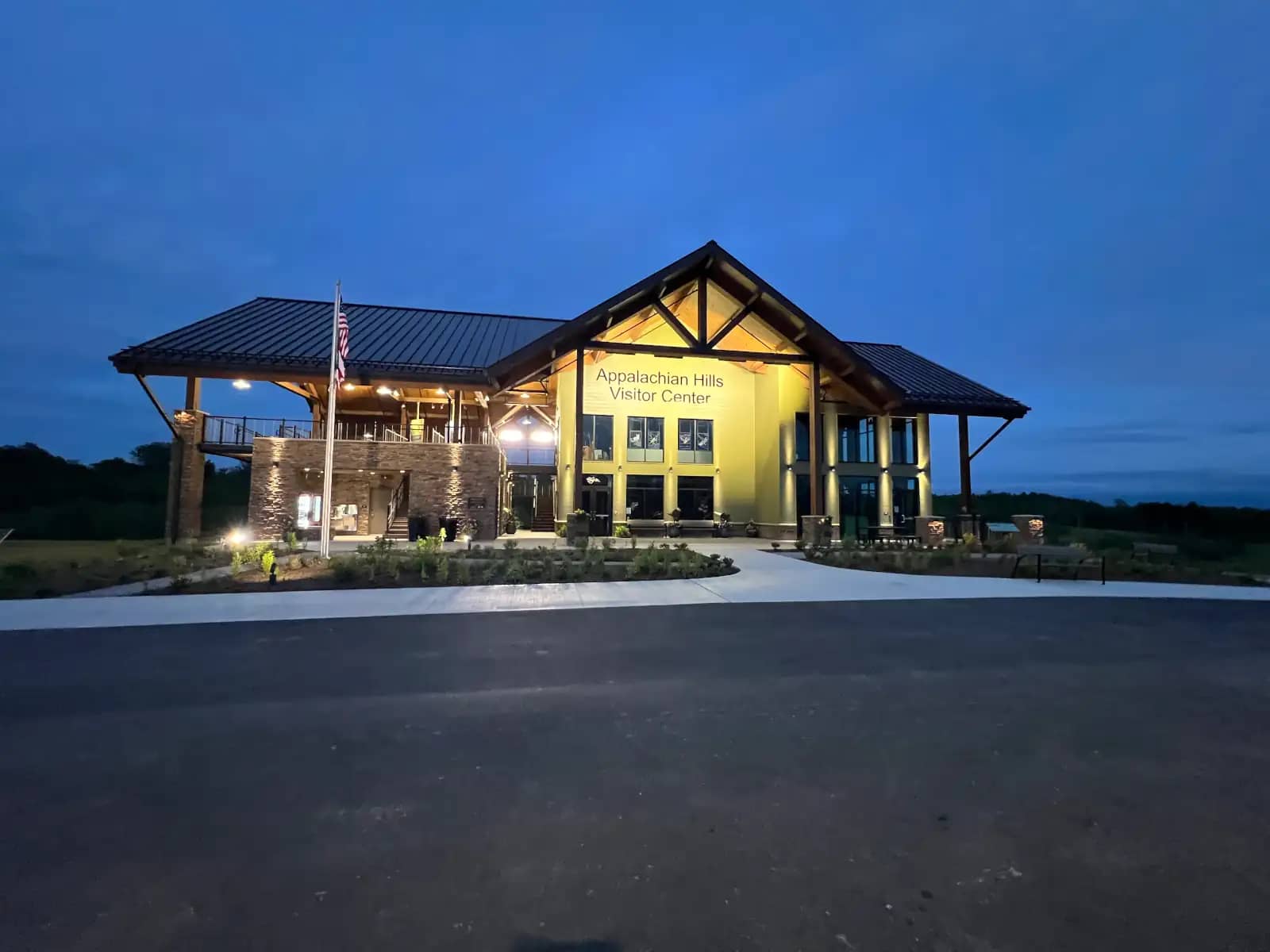 A wide exterior view of the two-story Appalachian Hills Visitor Center beautifully illuminated by warm yellow lights against a deep blue twilight sky.