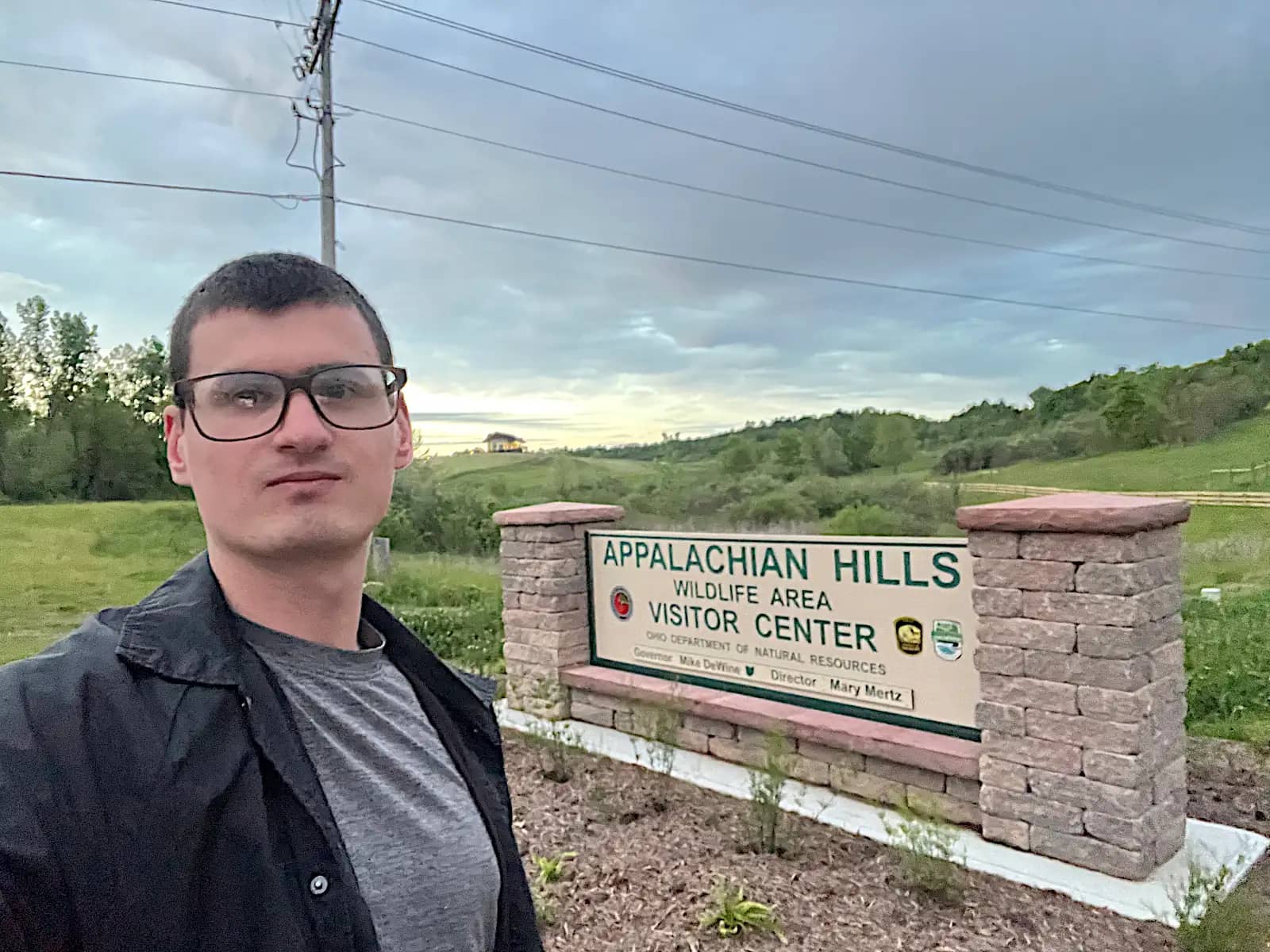 A close-up selfie of a man wearing glasses and a black jacket, standing in front of the stone-pillared Appalachian Hills Wildlife Area entrance sign.