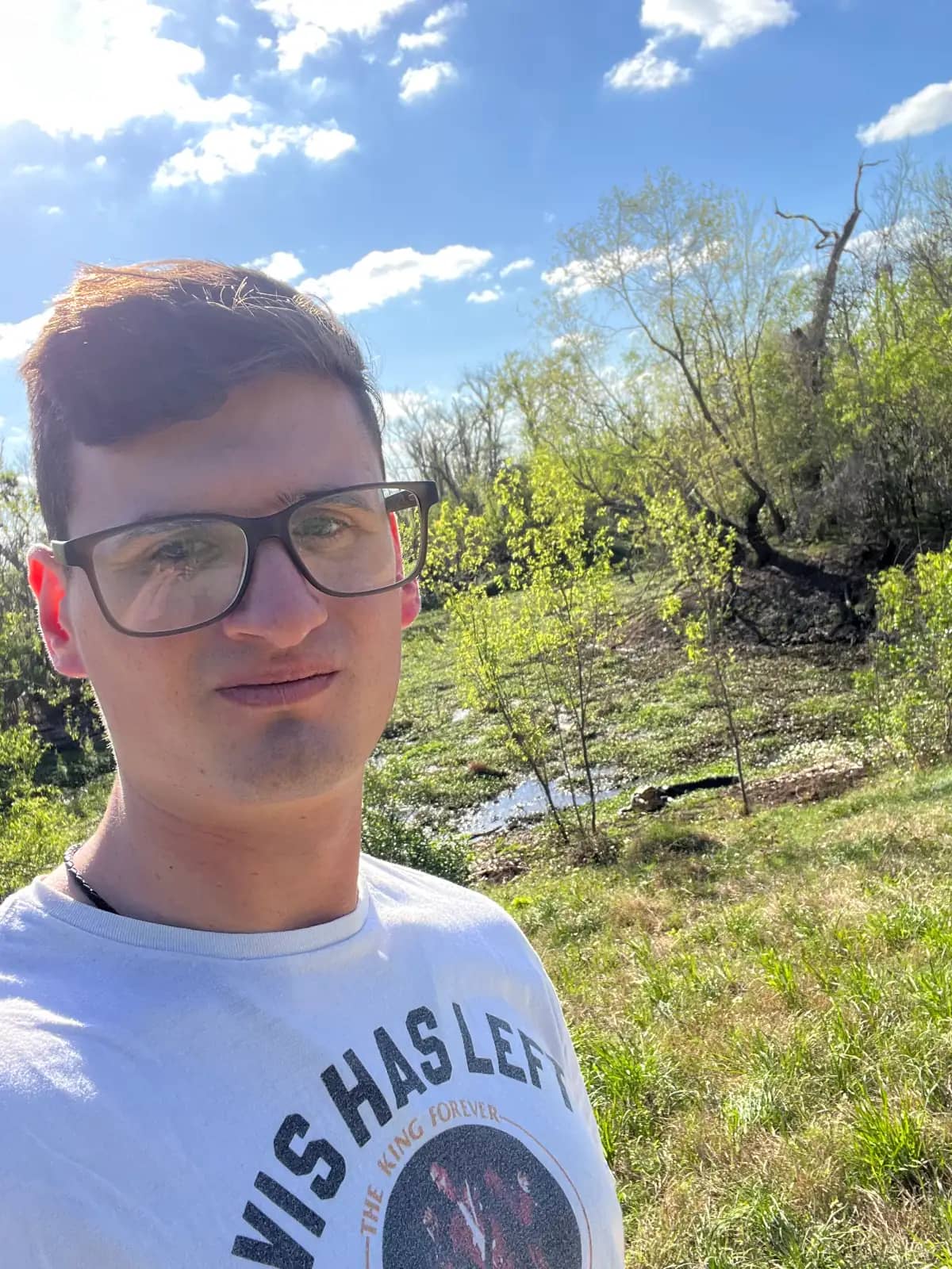 A selfie of a man with glasses and a white t-shirt with the flat, grassy marshlands of Brazos Bend State Park stretching out behind him with a giant alligator with it's mouth open in the background.