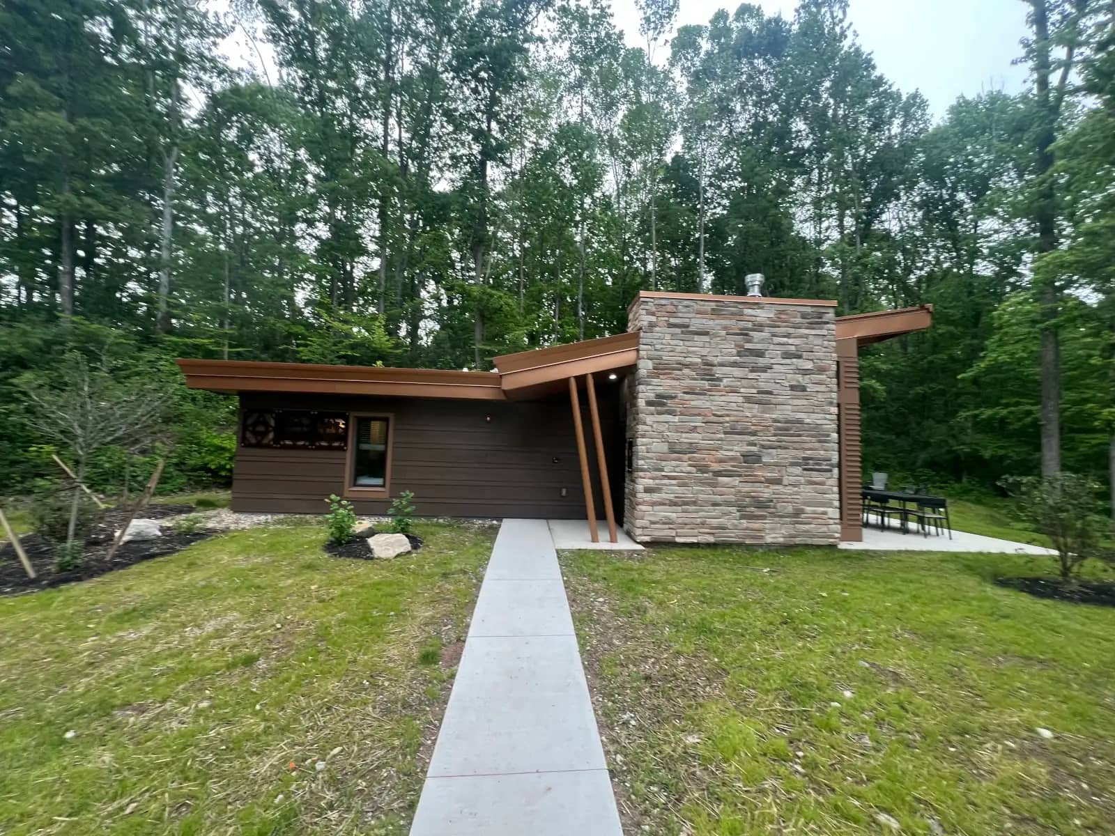 A wide exterior shot of a brand new, modern single-story cabin at Jesse Owens State Park featuring a prominent stone chimney, dark wood siding, and a clean concrete walkway.