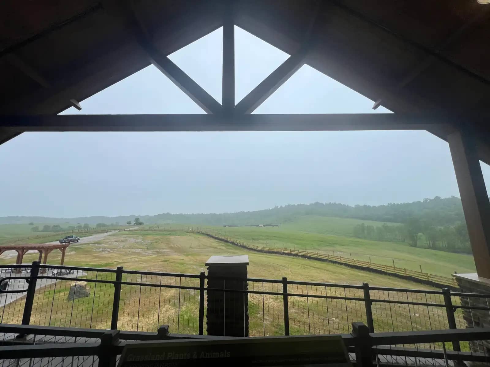 A view looking out from beneath the heavy wooden beams of a covered observation deck, showcasing a vast landscape of rolling green hills under a misty, overcast sky.