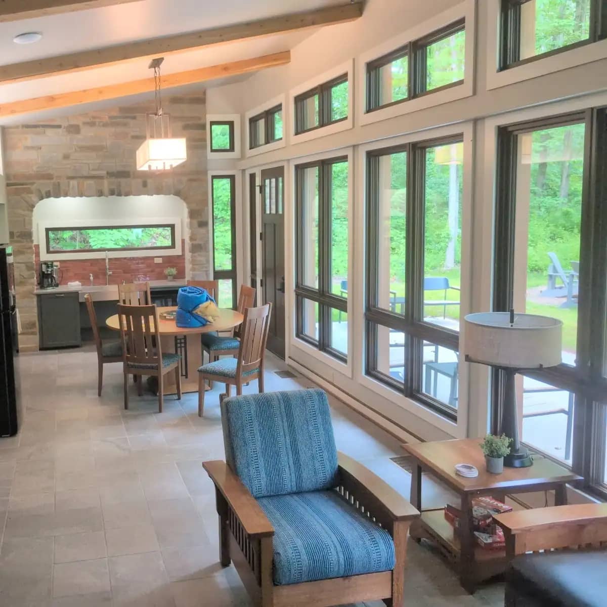 The sunlit interior of a modern cabin showing a wall of large windows looking out into green woods, a blue patterned armchair, a wooden dining table, and a stone accent wall in the kitchen area.