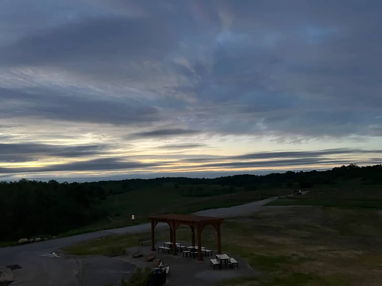An elevated dusk shot looking down at an empty picnic shelter and a road winding through rolling green hills under a dramatic, cloudy sunset sky.