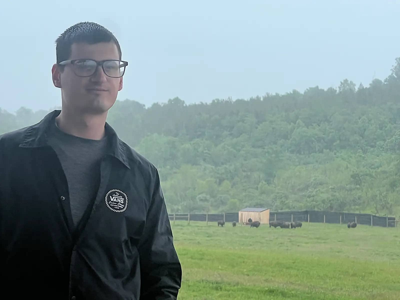 A man in glasses and a dark jacket standing on an outdoor observation deck, with a herd of distant buffalo visible grazing in a fenced green pasture behind him.