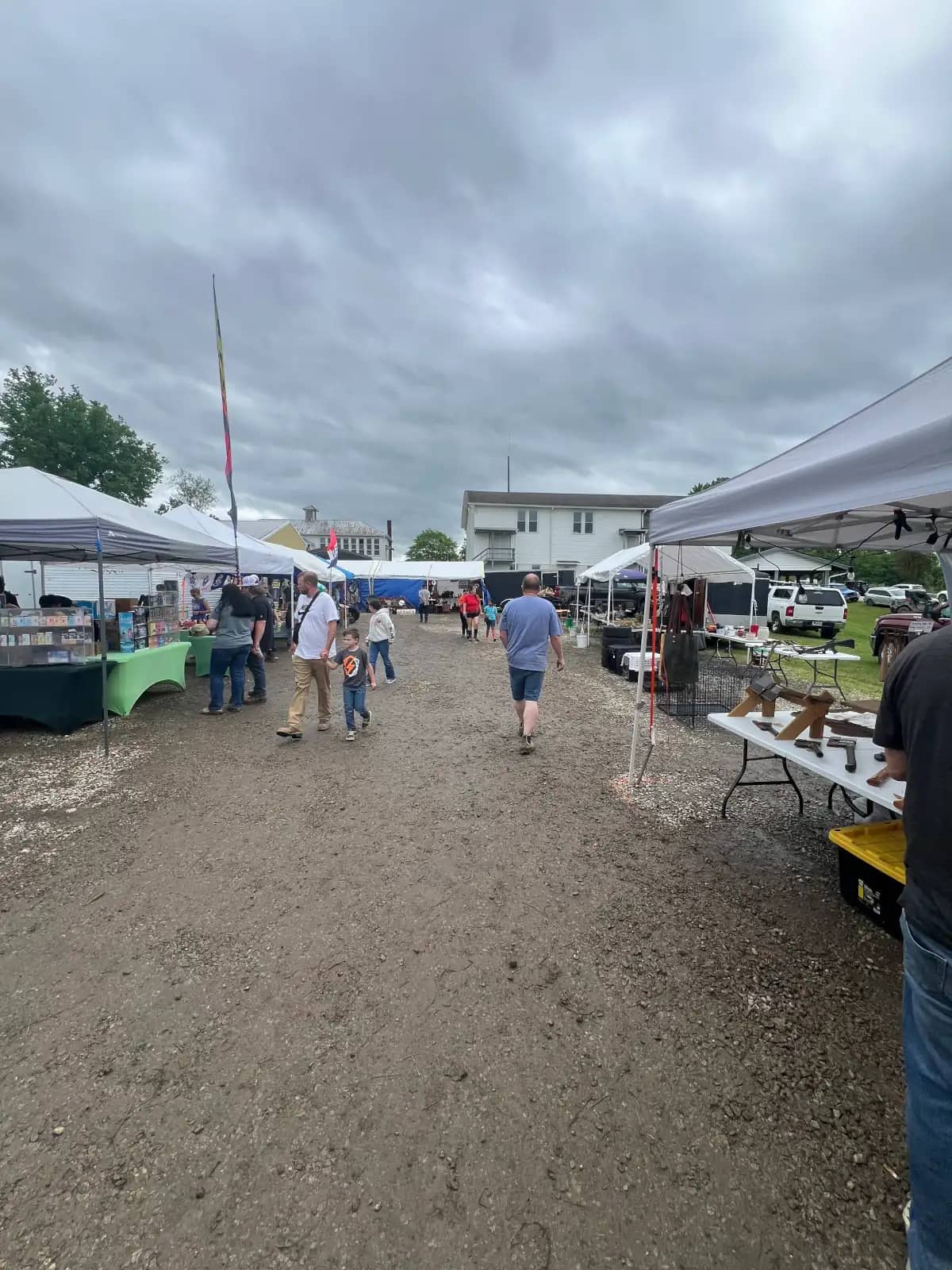 A wide view of an outdoor flea market under a gloomy sky, with visitors walking down a dirt path lined with white pop-up tents and vendor tables.