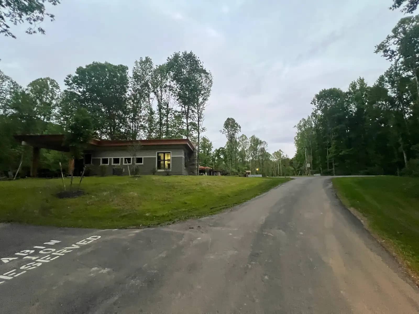 A paved road leading past a modern cabi, surrounded by green lawns and dense woods under a cloudy sky.
