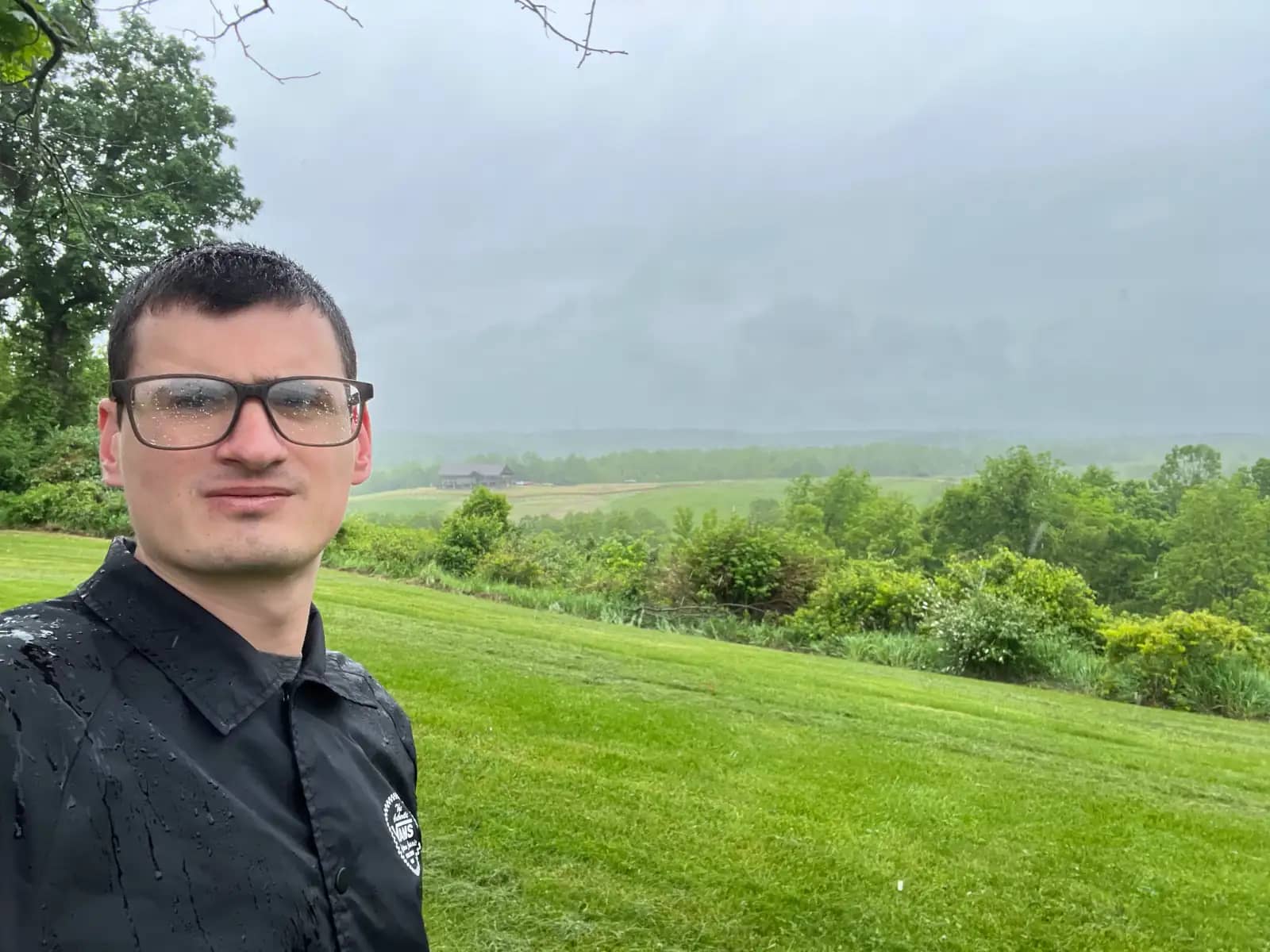 A rain-soaked selfie of a man with visible water droplets on his glasses and black jacket, standing on a vibrant green hillside overlooking a foggy, overcast valley.