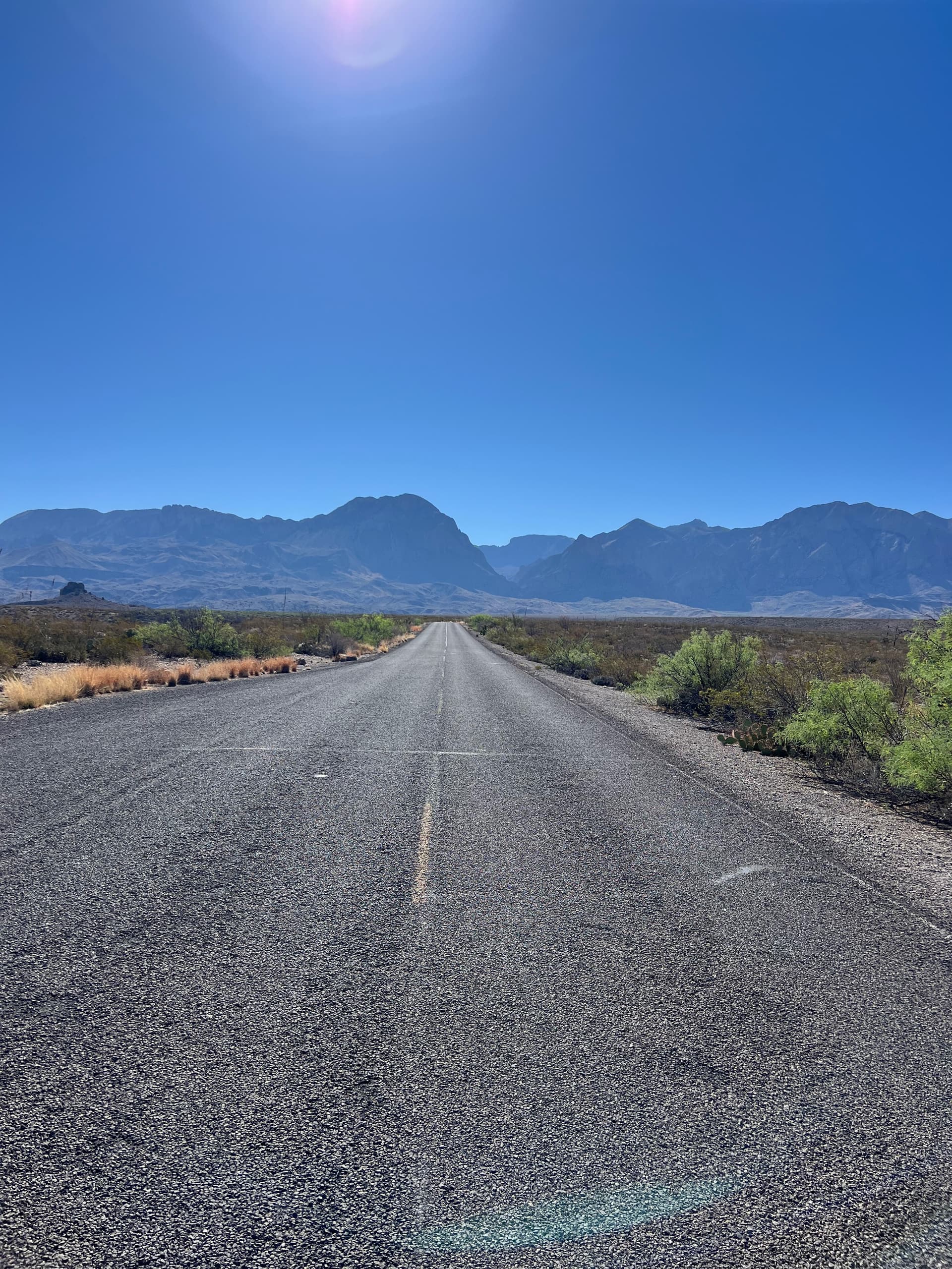 A straight paved road stretching toward a massive blue mountain range under a bright, cloudless sky.