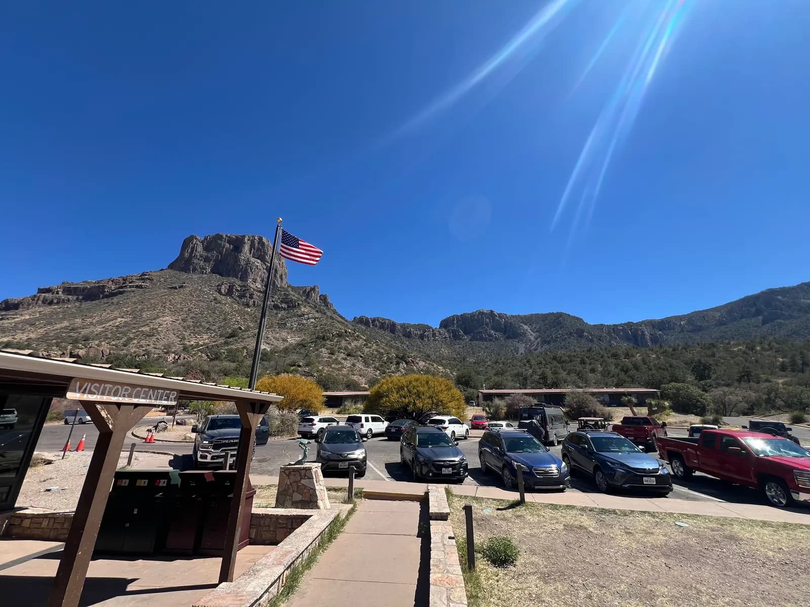 A parking lot filled with cars in front of a visitor center building, with a massive flat-topped mountain peak rising behind it.