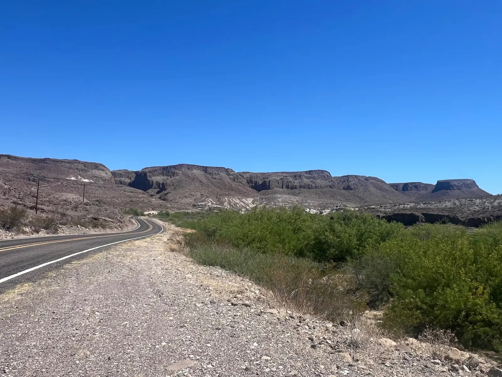 A paved highway curves through a desert landscape with vibrant green bushes on the right and tall, flat-topped mountains in the distance under a clear blue sky.