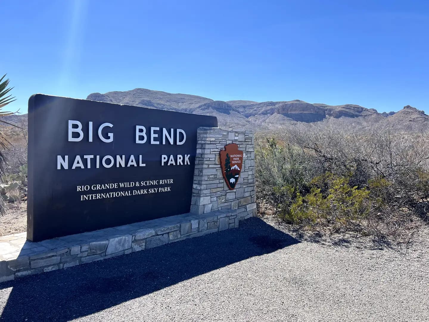 A large, modern dark metal sign on a small stone base that reads "BIG BEND NATIONAL PARK," with the NPS arrowhead logo. Mountainous landscape is in the background.