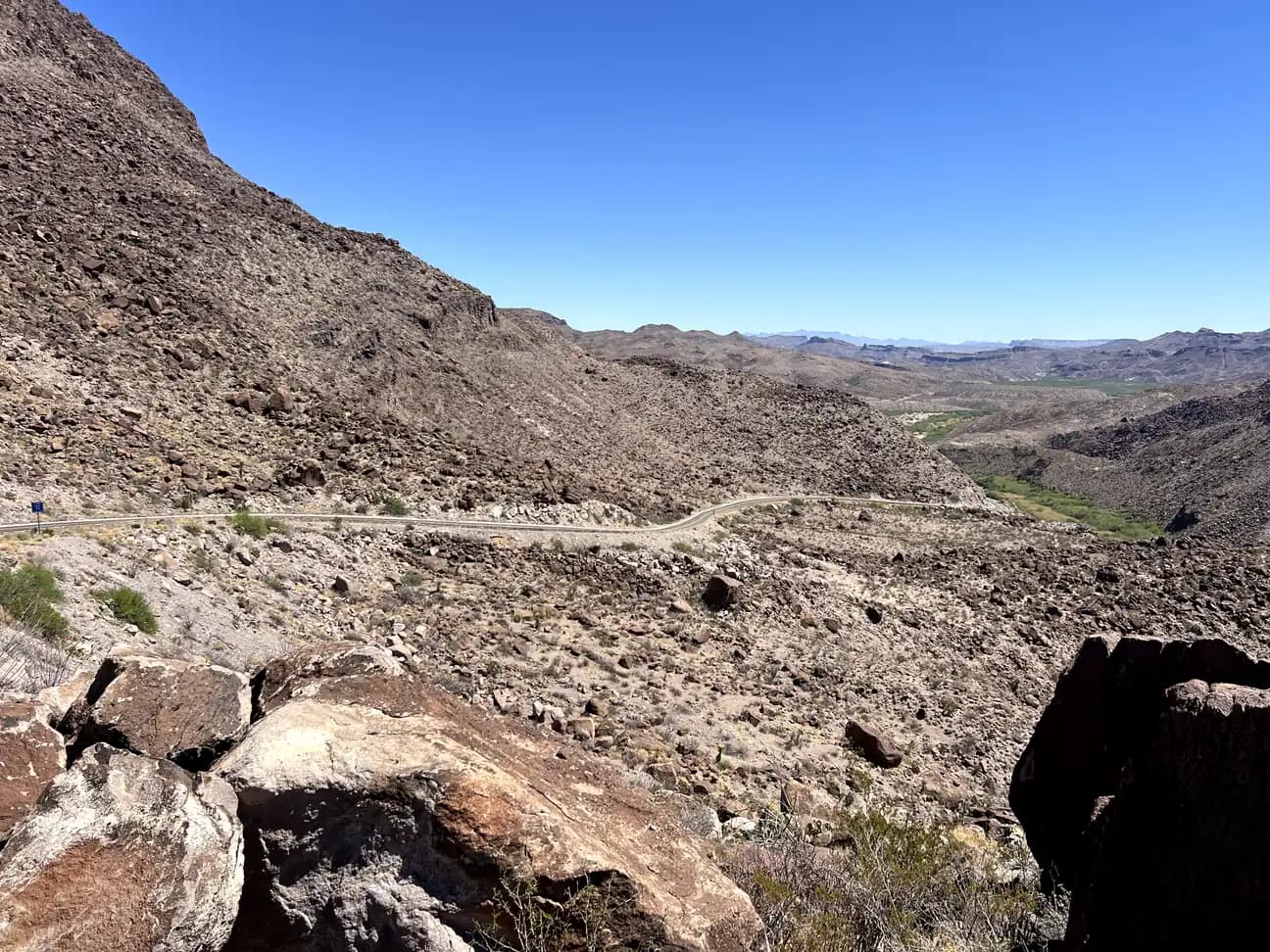 A high-angle view of a winding two-lane highway carving through a steep, rocky mountain slope under a clear blue sky.