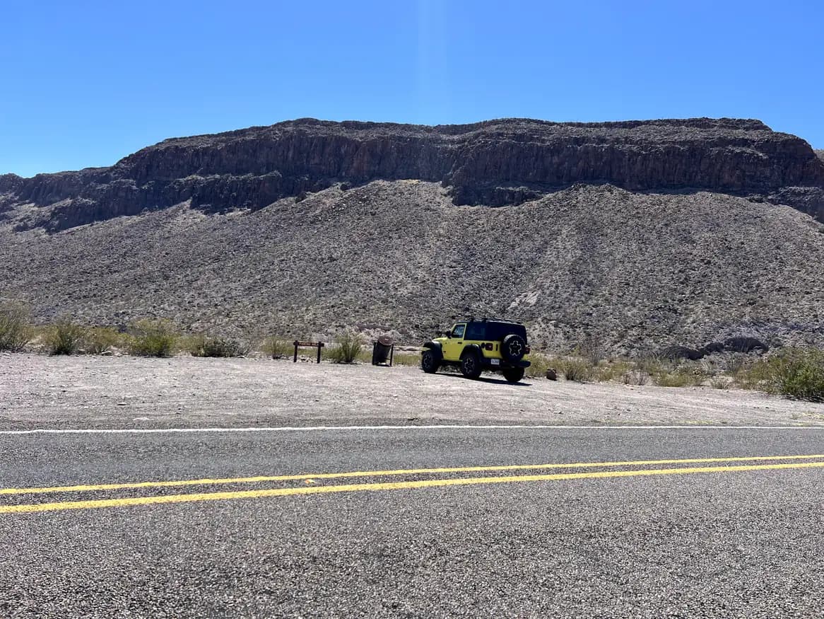A bright yellow Jeep Wrangler parked on a gravel roadside at the base of a massive, dark brown rocky mountain under a clear blue sky.