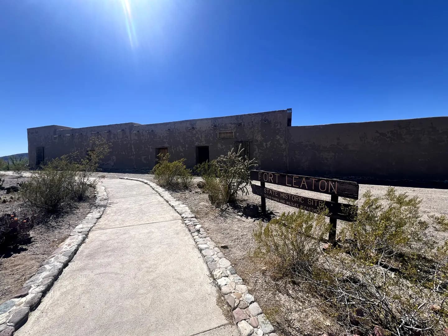 A concrete path lined with stones leads toward a long, brown adobe building under a bright sun. A wooden sign in the foreground reads "Fort Leaton."