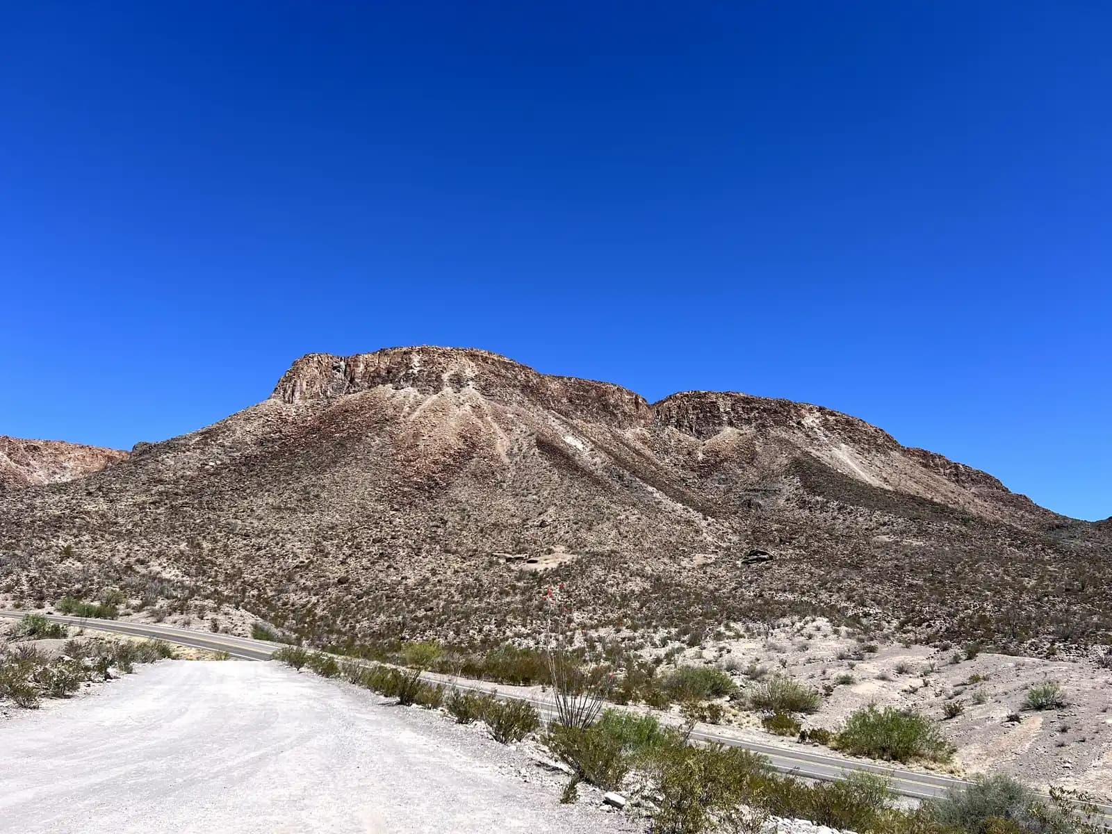 A wide shot of a winding desert road leading toward a massive, light-colored mountain with visible geological layers and a vast blue horizon.