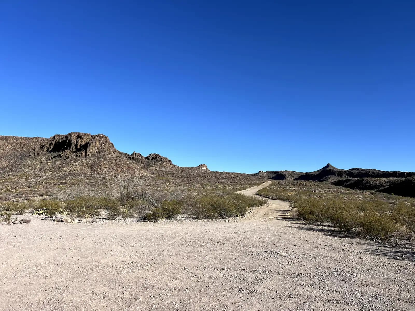 A winding dirt and gravel road leads into a desert landscape with rocky hills and sparse green scrub under a vibrant, cloudless blue sky.