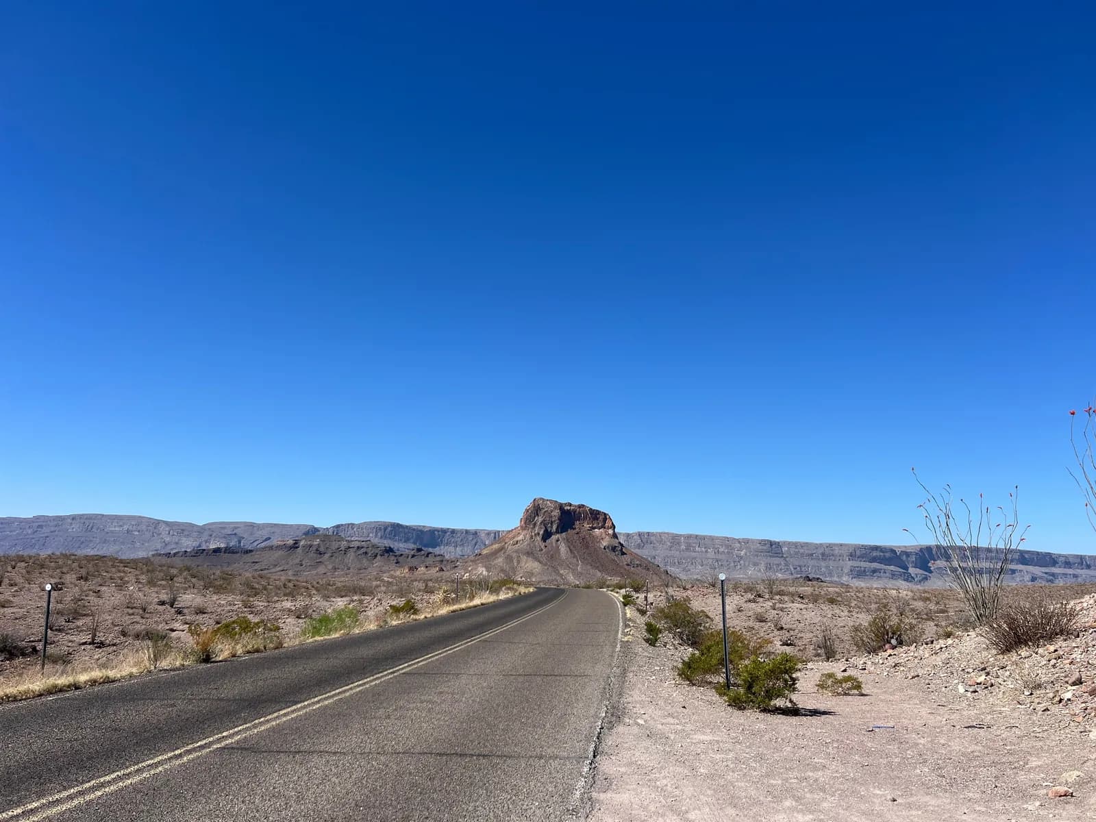 A long road leads toward a prominent, solitary volcanic butte under a deep blue desert sky.