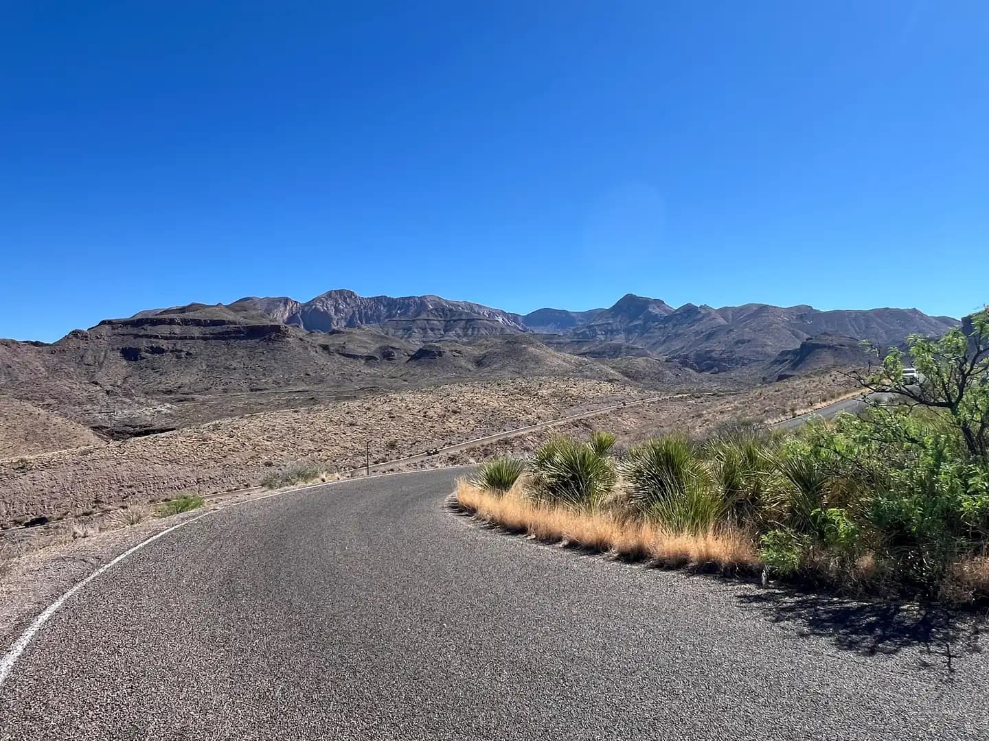 A paved road curves sharply through a dry desert landscape toward distant, layered mountain peaks.