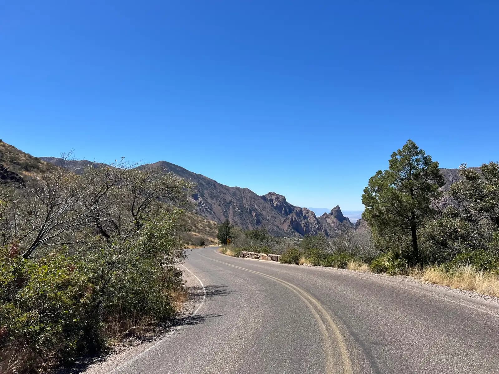 A two-lane paved road curves through a desert landscape toward high, rugged mountains under a clear blue sky.
