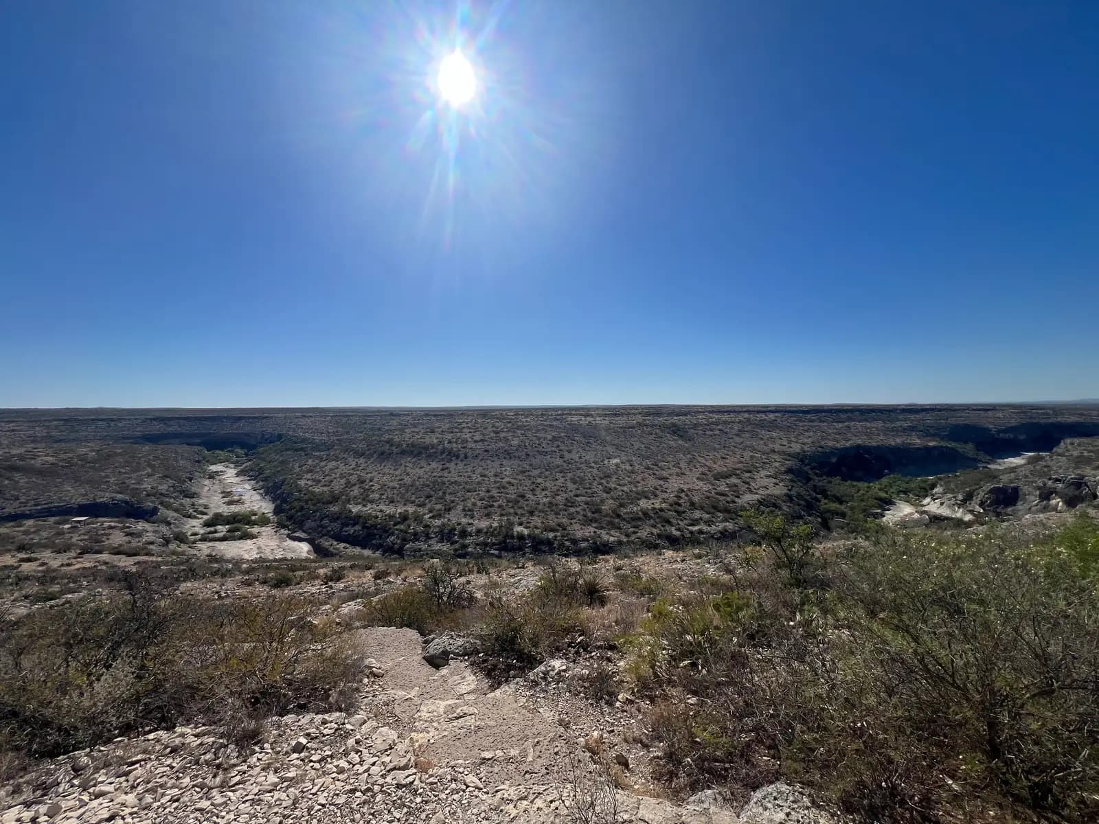 An expansive view of a flat, arid landscape with a canyon winding to the right under a bright, high sun.