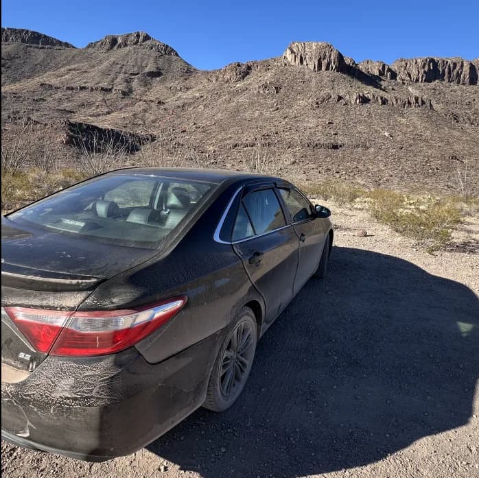 A dusty black Toyota Camry parked at a gravel trailhead, with steep, rocky desert mountains rising sharply in the background under a clear sky.