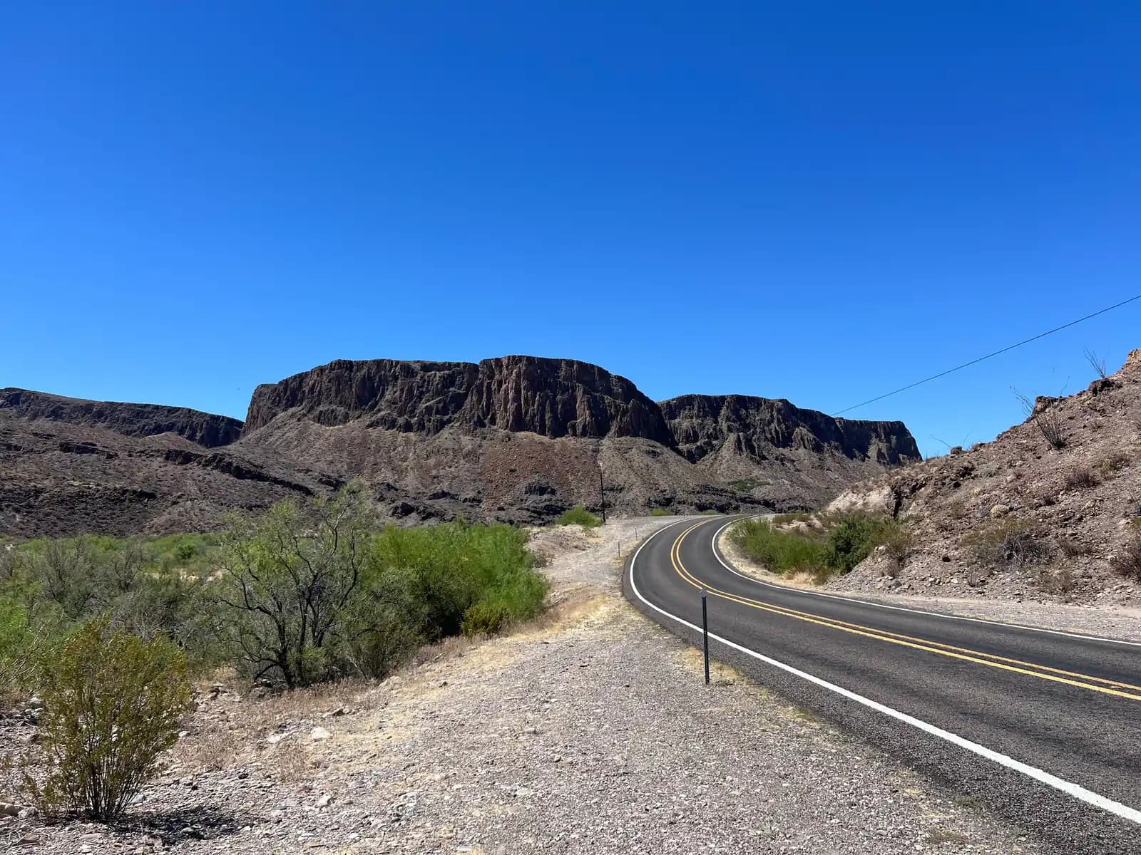 A view of a winding asphalt desert highway (River Road) with yellow lines, cutting through arid, rocky terrain under a vibrant blue sky toward dark, flat-topped mesas.