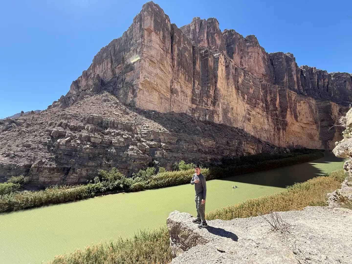 A man stands on a rocky ledge overlooking a wide green river with massive limestone cliffs rising above.