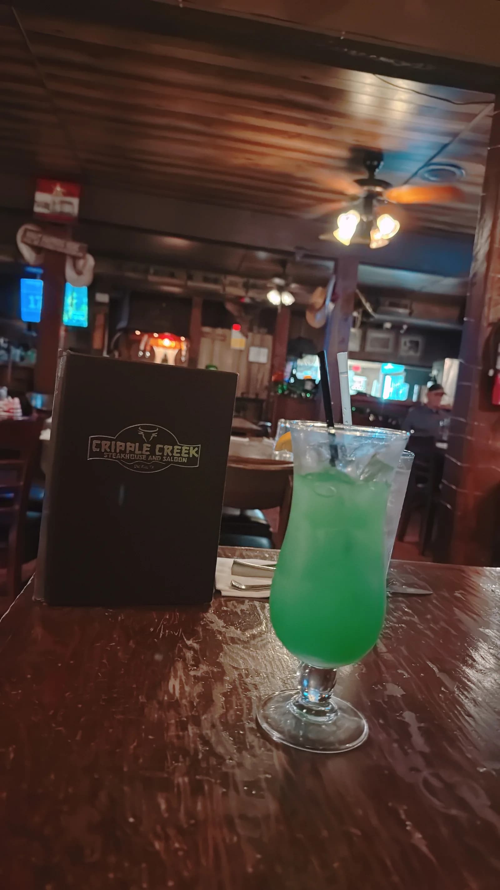 A close-up of a vibrant green cocktail with two straws on a wooden table. A dark Cripple Creek Steakhouse and Saloon menu sits in the background.