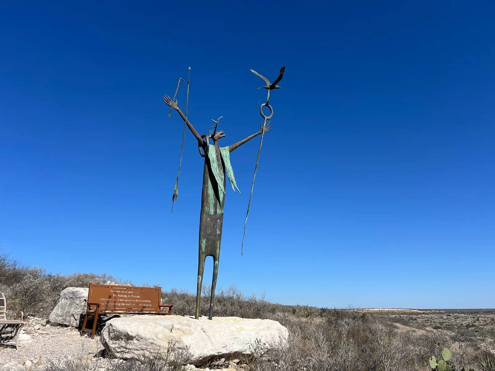 A tall, slender bronze sculpture of a figure with raised arms on a large rock base in front of a blue sky.