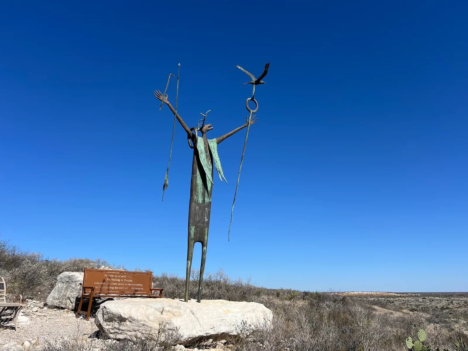 A tall, slender bronze sculpture of a figure with raised arms on a large rock base in front of a blue sky.