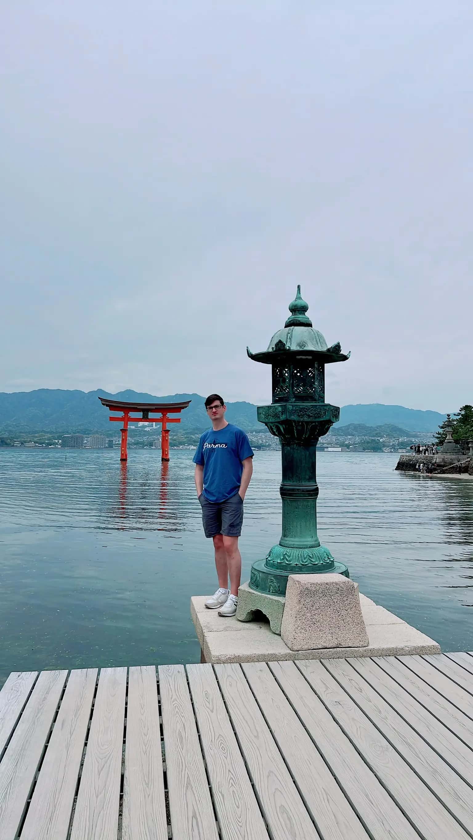 The Lost Shepard standing beside a stone lantern on Miyajima Island with the iconic floating torii gate of Itsukushima Shrine and forested mountains in the background