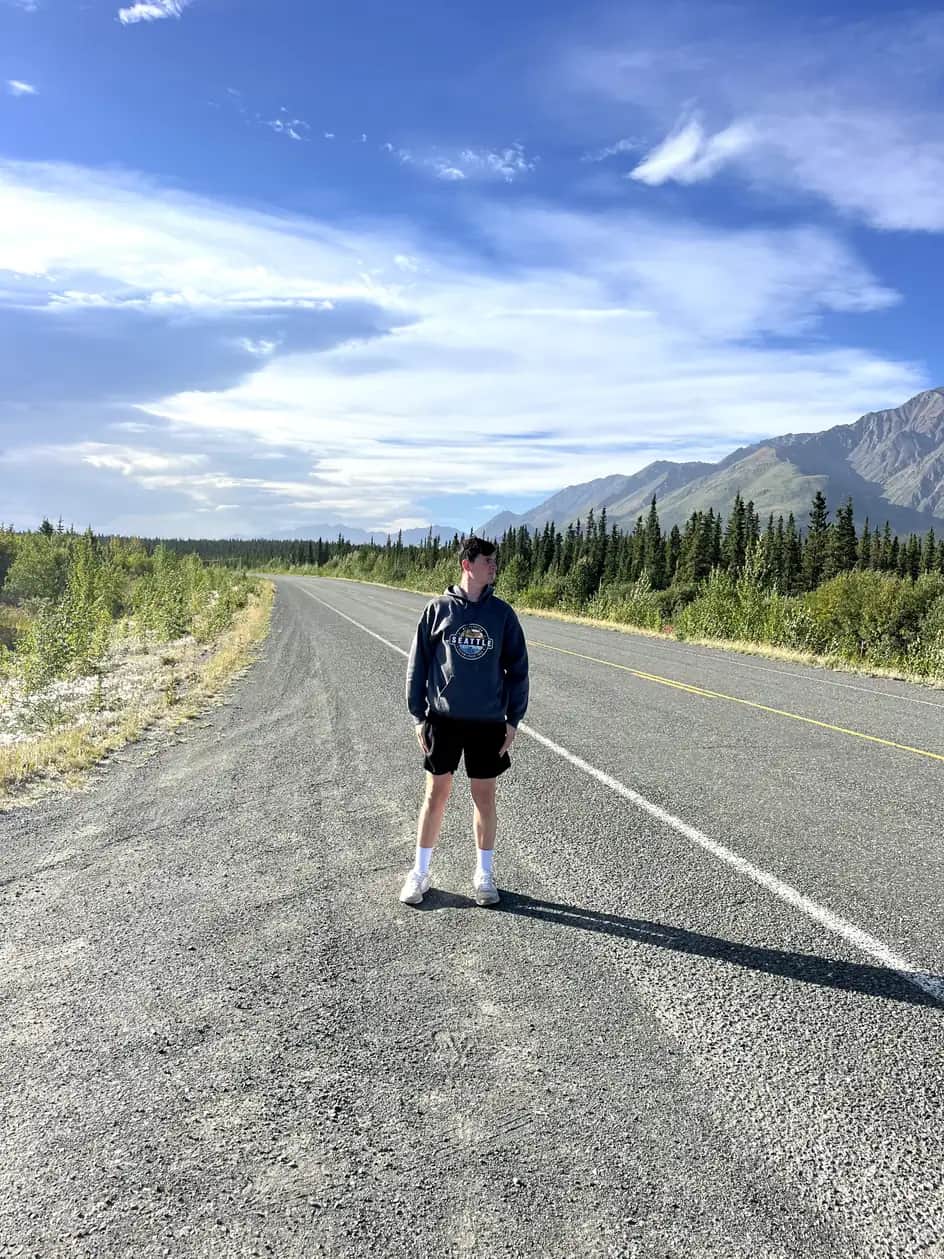 The Lost Shepard standing in the middle of the Alaska Highway with dense boreal forest and distant mountains under a partly cloudy sky