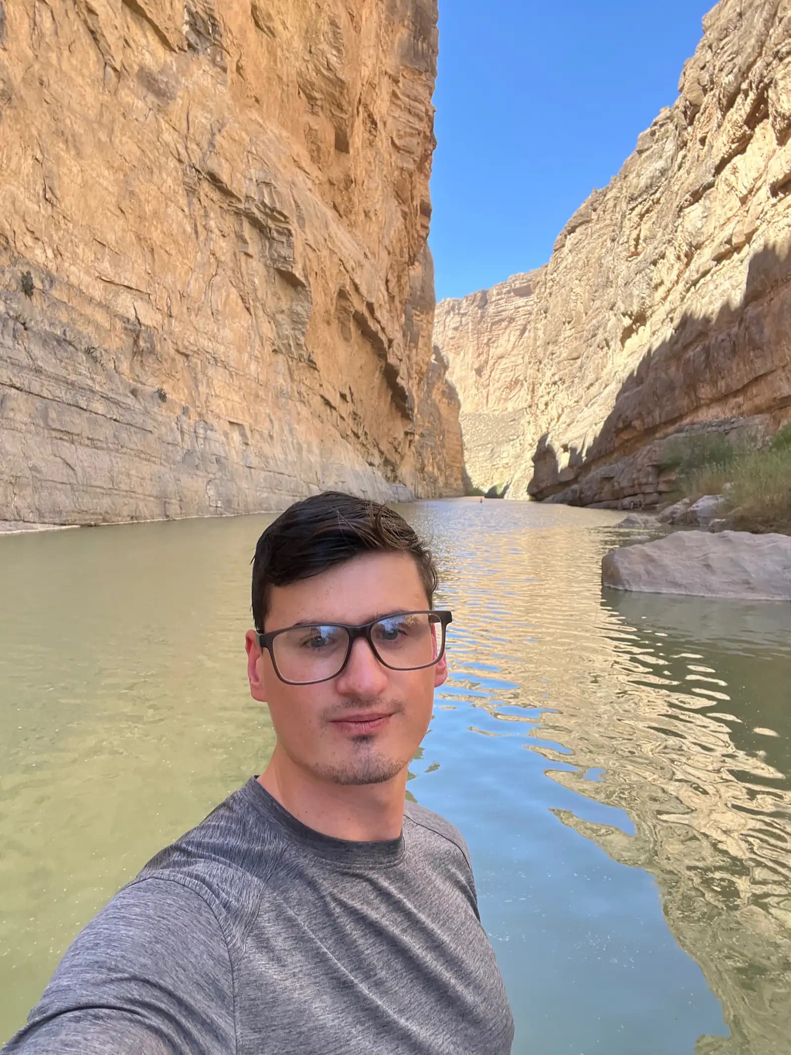 A selfie of a man in glasses standing at the edge of a river inside a narrow canyon with tall, sunlit rock walls.