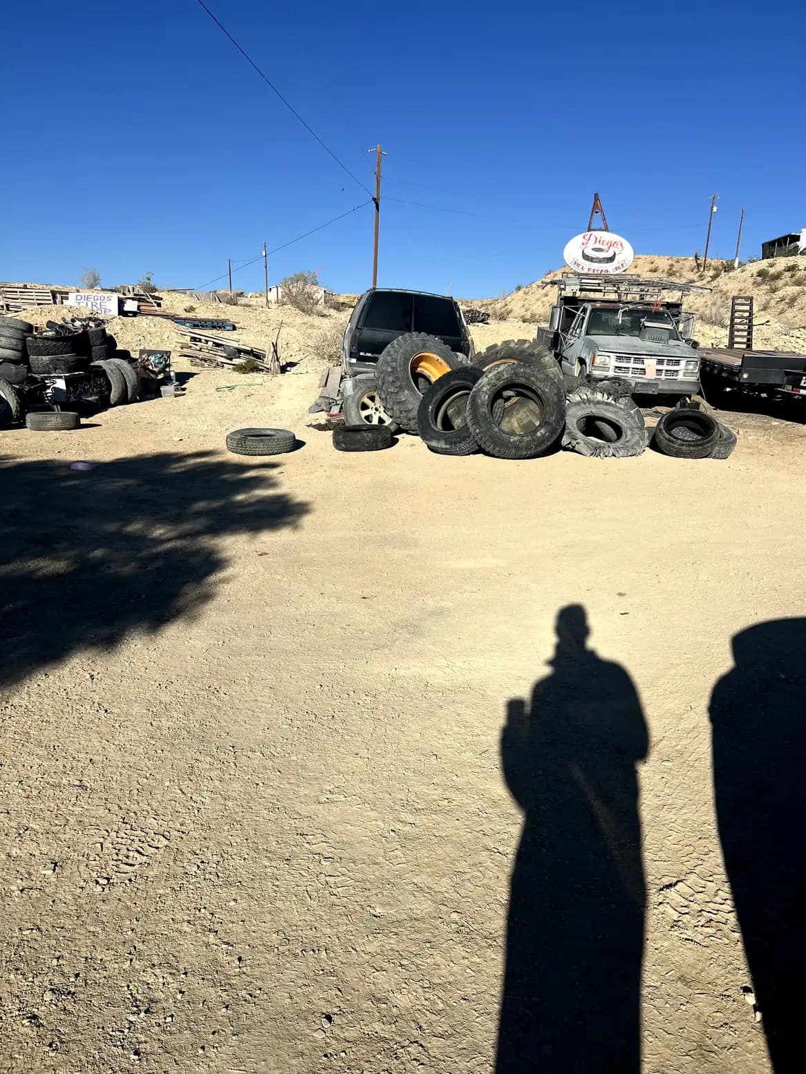 A desert tire repair shop featuring piles of various sized tires, old trucks, and a "Diego's" sign against a sandy hill and bright blue sky.