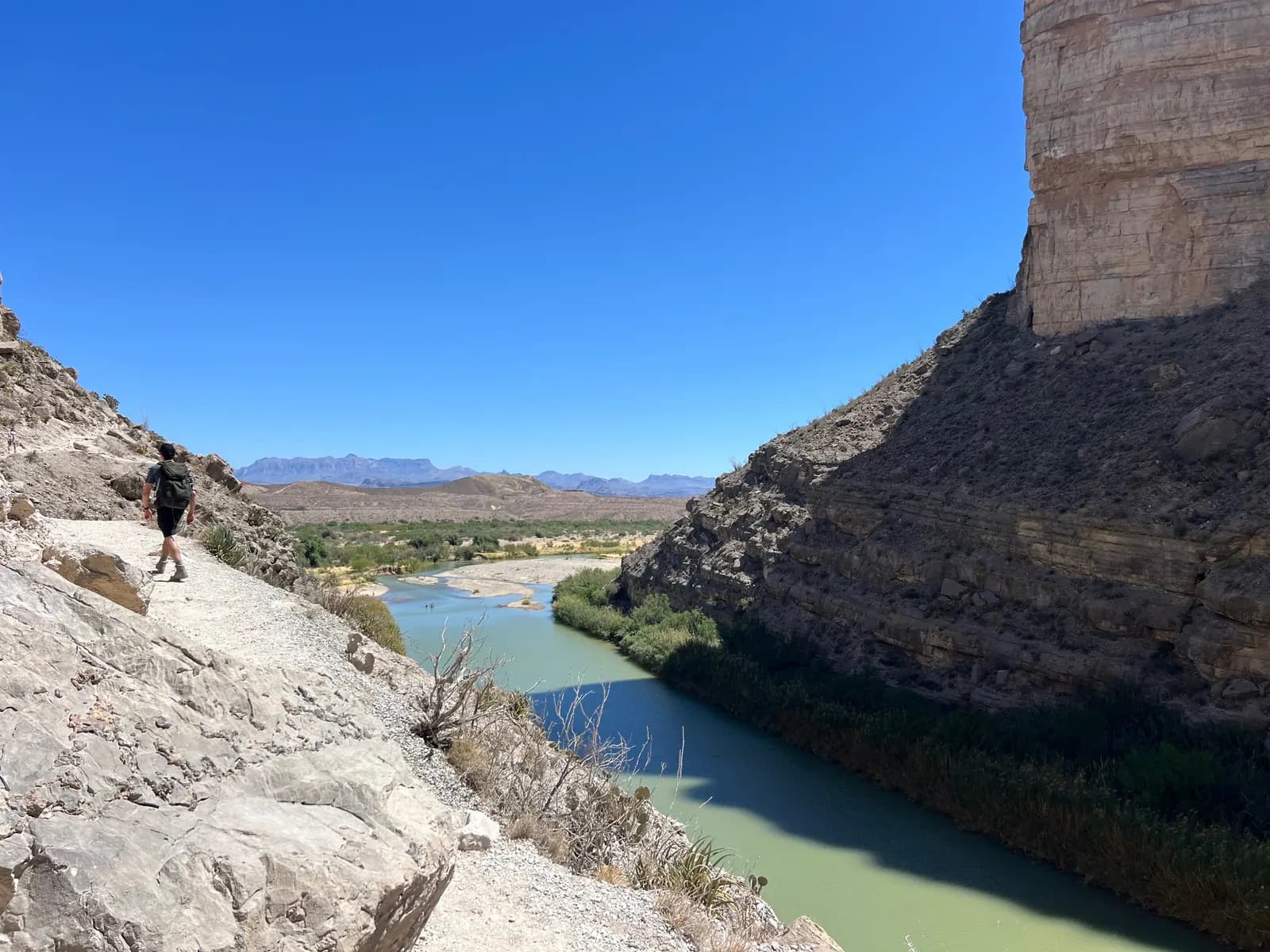 A hiker walks along a narrow, rocky cliffside trail overlooking a wide green river at the bottom of a canyon.