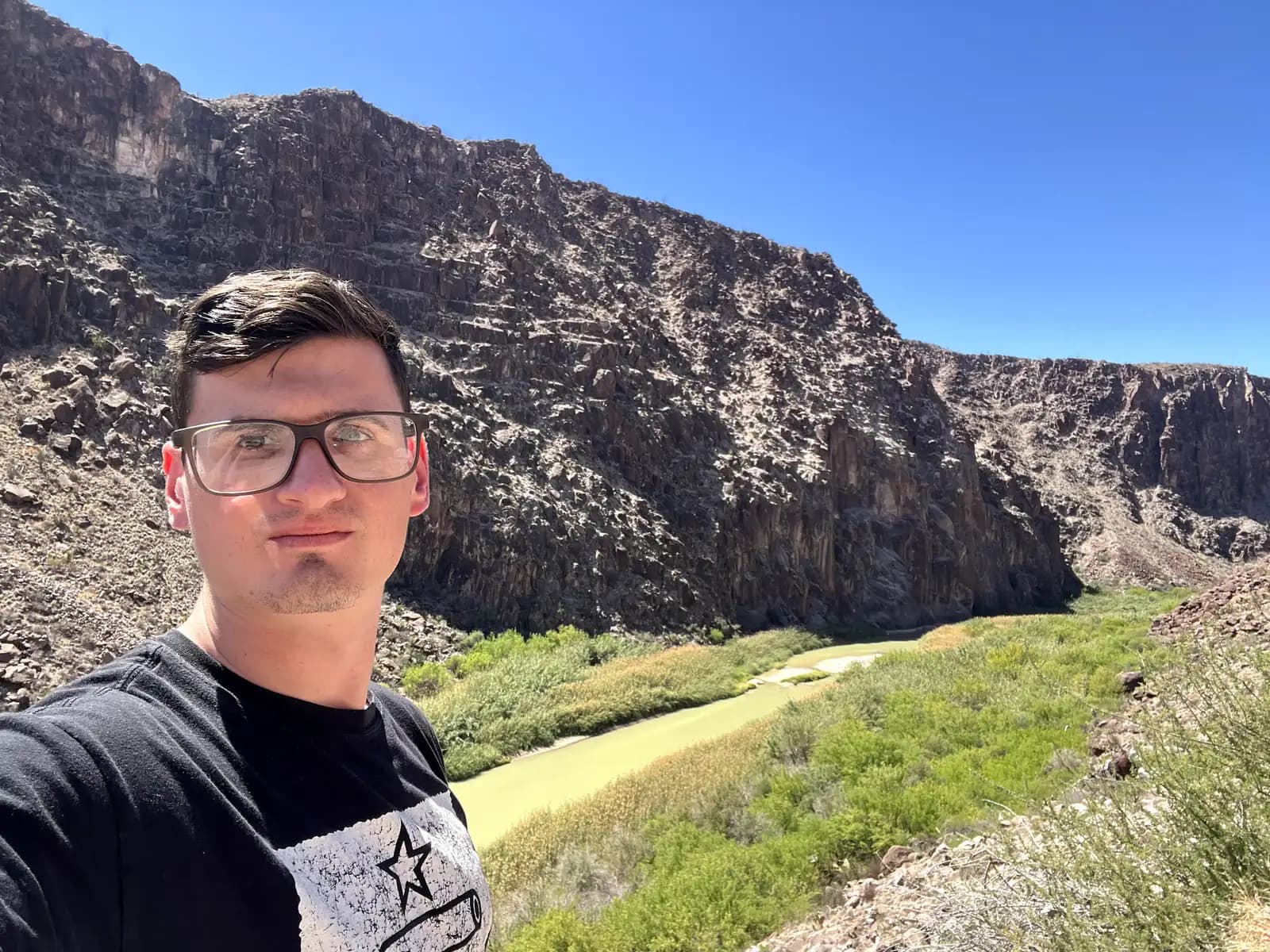 A selfie of a man with glasses and a black shirt in front of the winding Rio Grande and the large, rocky mountains of Mexico.