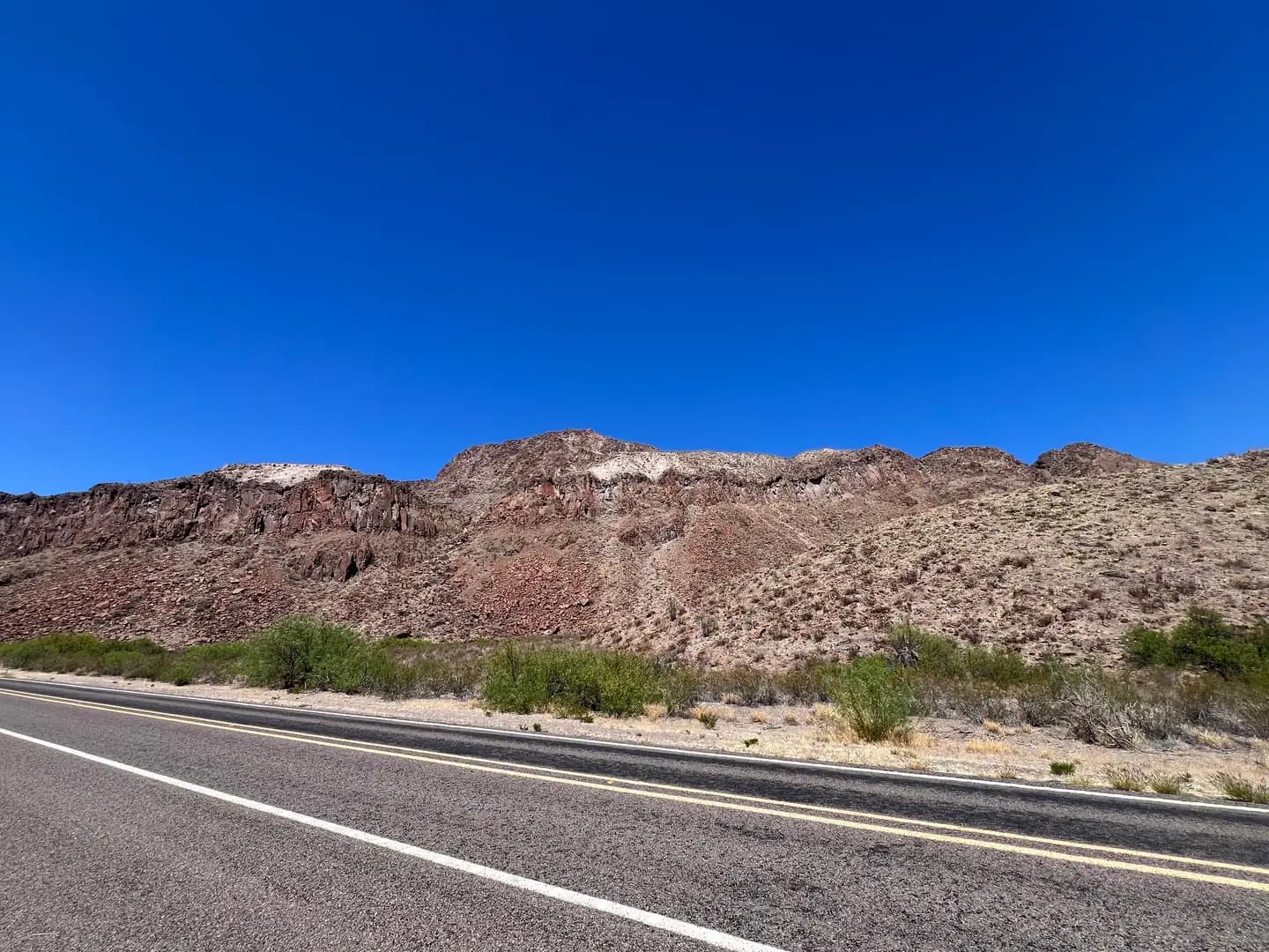 An asphalt road with yellow lines stretches past dark, reddish volcanic rock mountains and desert greenery under a deep blue sky.