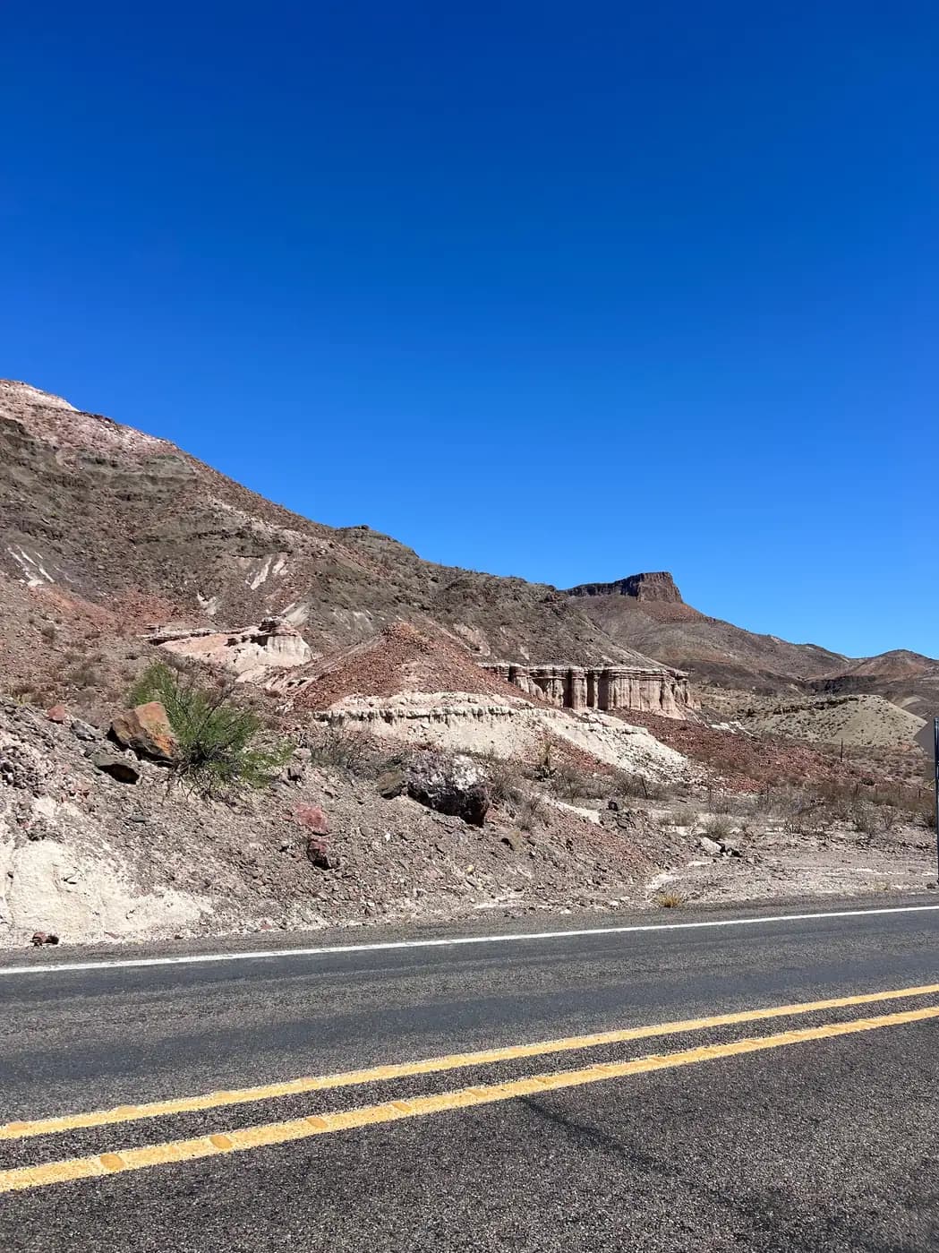 A roadside view of steep desert mountains featuring distinct white and reddish-brown rock layers and jagged formations under a clear sky.