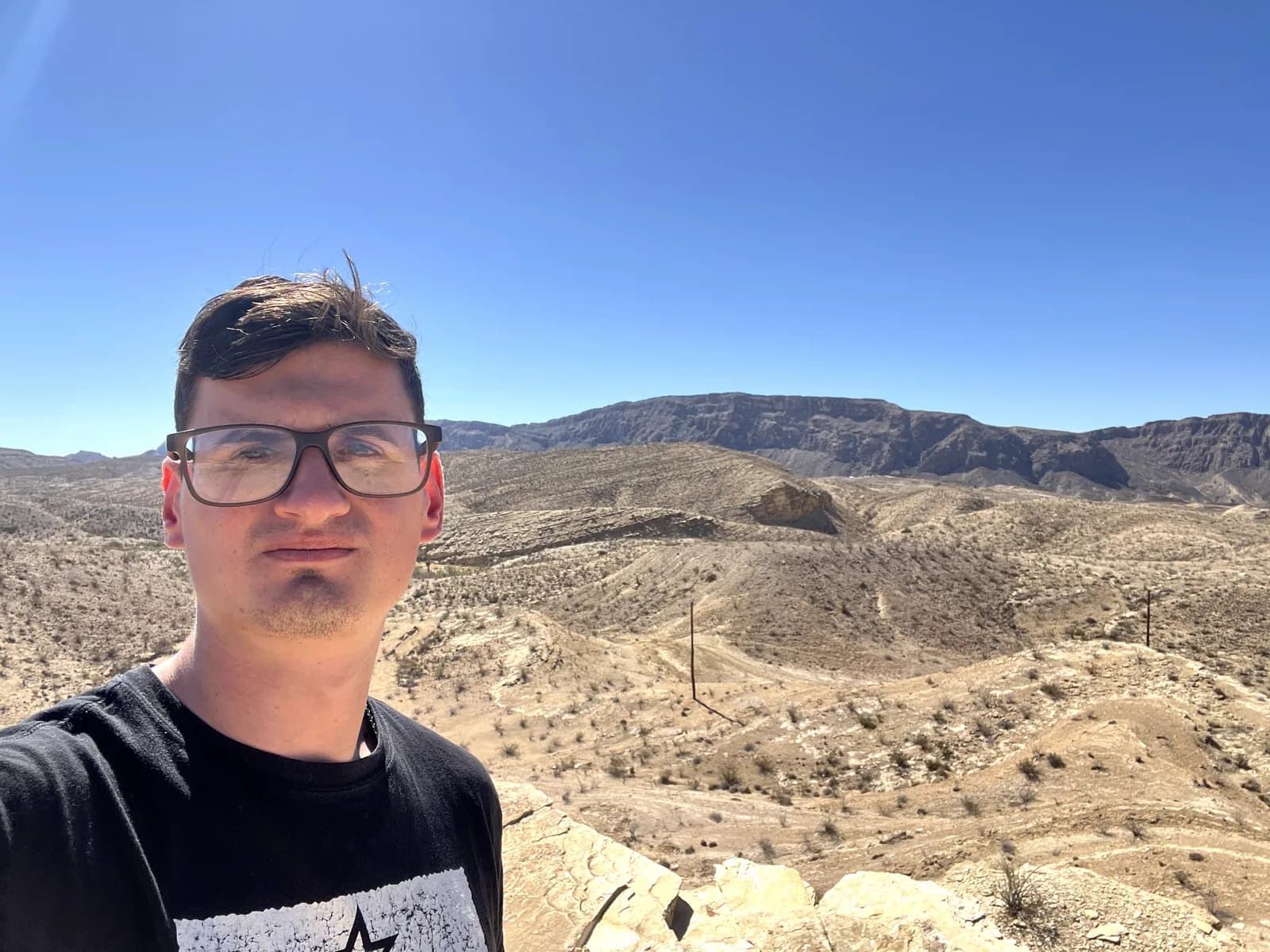 A selfie of a man with glasses in front of an expansive desert valley with rolling hills and a large, flat-topped mountain under a bright blue sky.