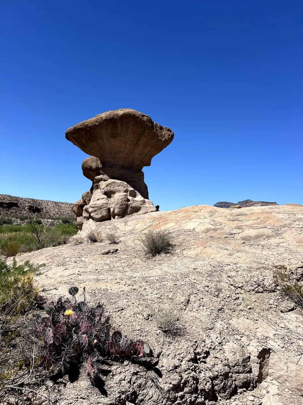 A large, mushroom-shaped rock formation known as a hoodoo stands tall against a clear, deep blue sky in a desert landscape.