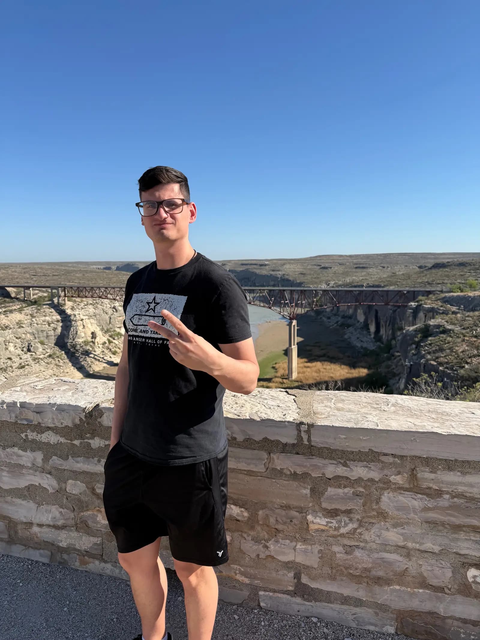 A portrait of a man in black clothing posing with a peace sign, with the Pecos River High Bridge in the background.