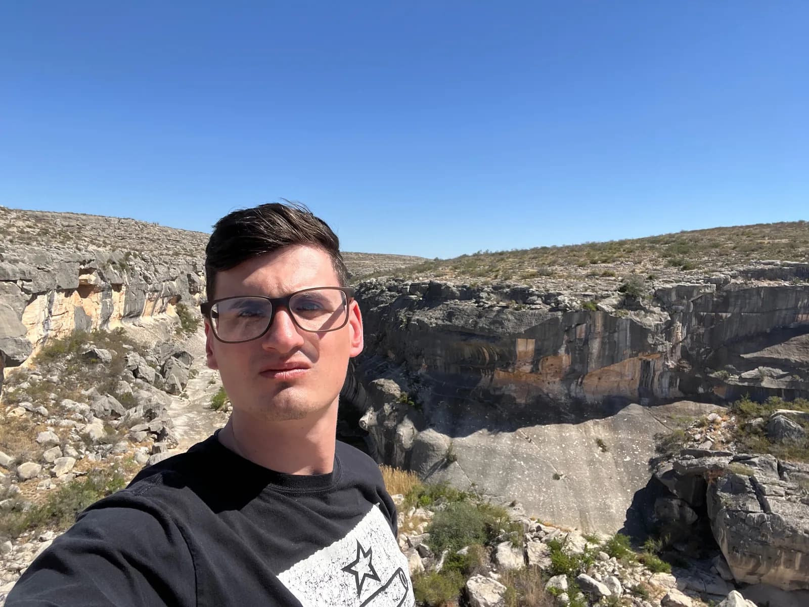 A low-angle self-portrait of a man with glasses against a backdrop of steep, rugged, multi-colored canyon walls under a clear blue sky.