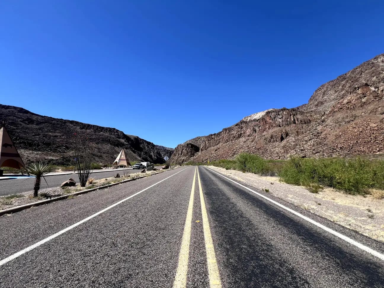 A low-angle view of a paved two-lane highway stretching toward a canyon. To the left, there are teepee-shaped rest areas under a bright blue sky.
