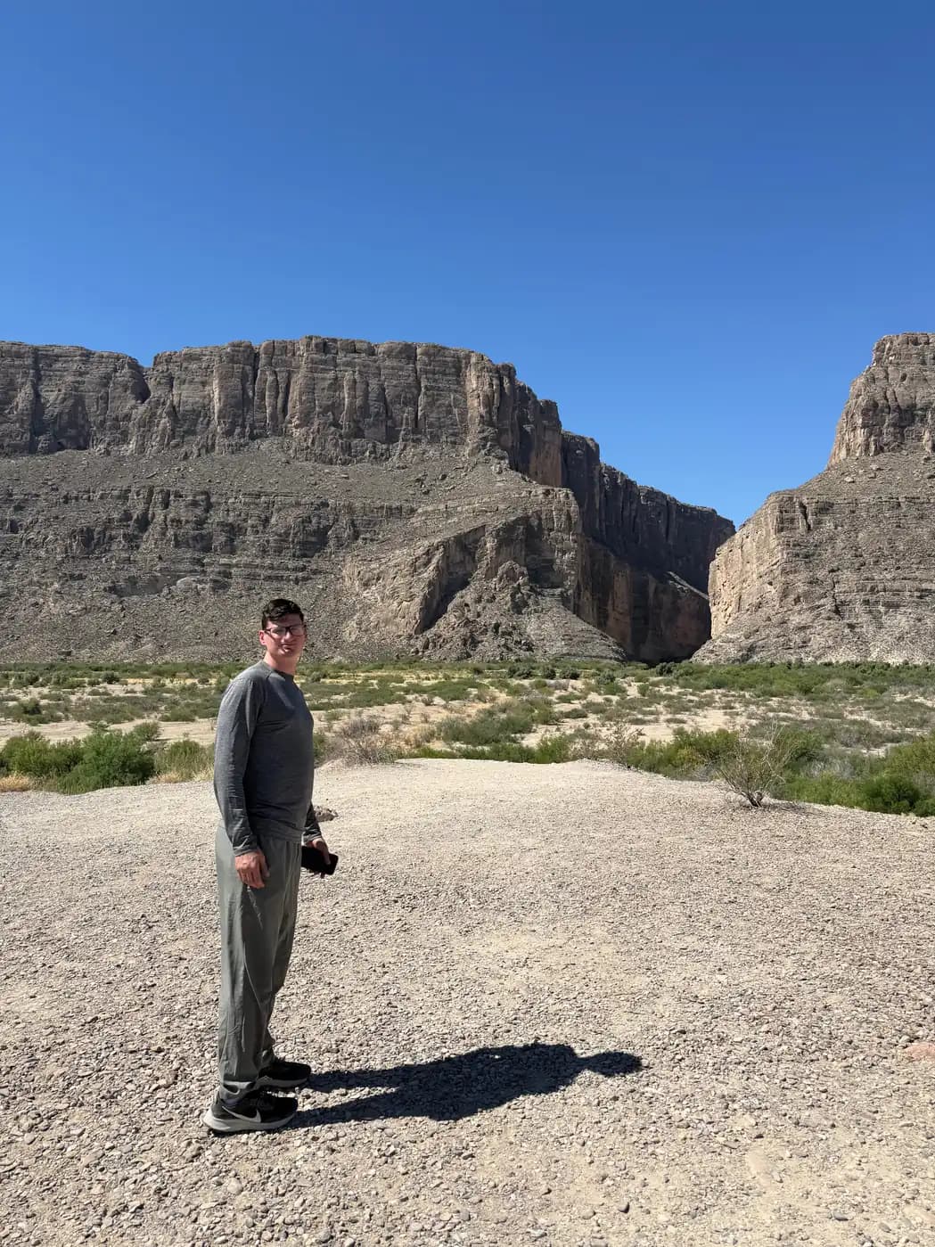 A man in a long-sleeve gray shirt stands in a rocky, open desert area with the massive cliffs of a canyon in the background.