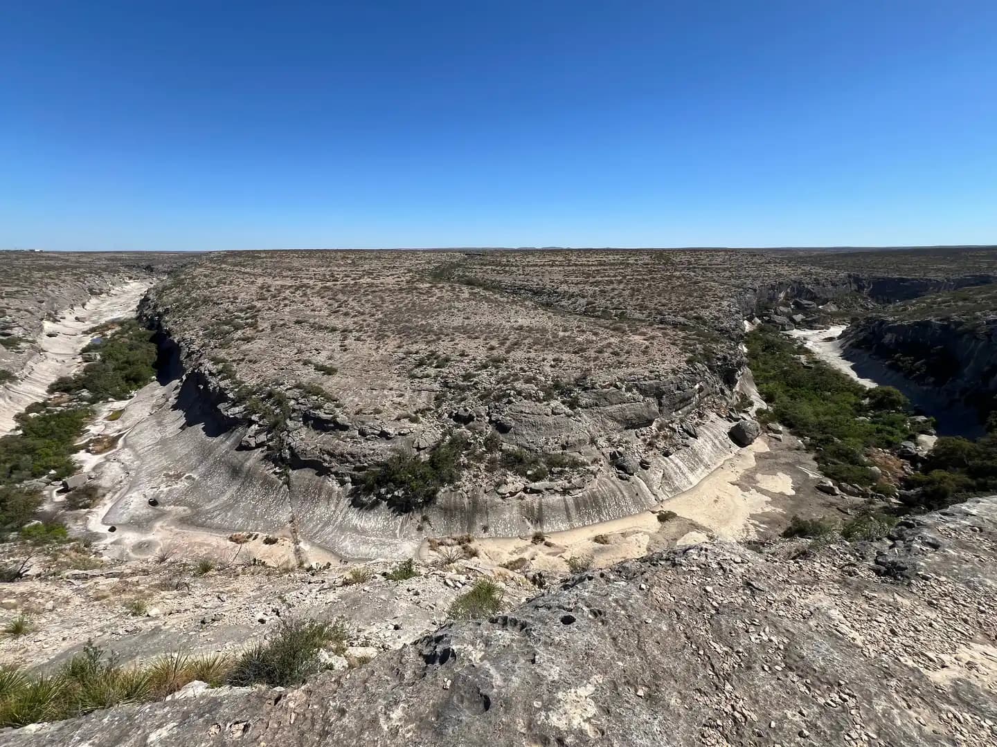 An elevated viewpoint of a wide, looping horseshoe bend in the desert canyon with dry riverbed and brush.