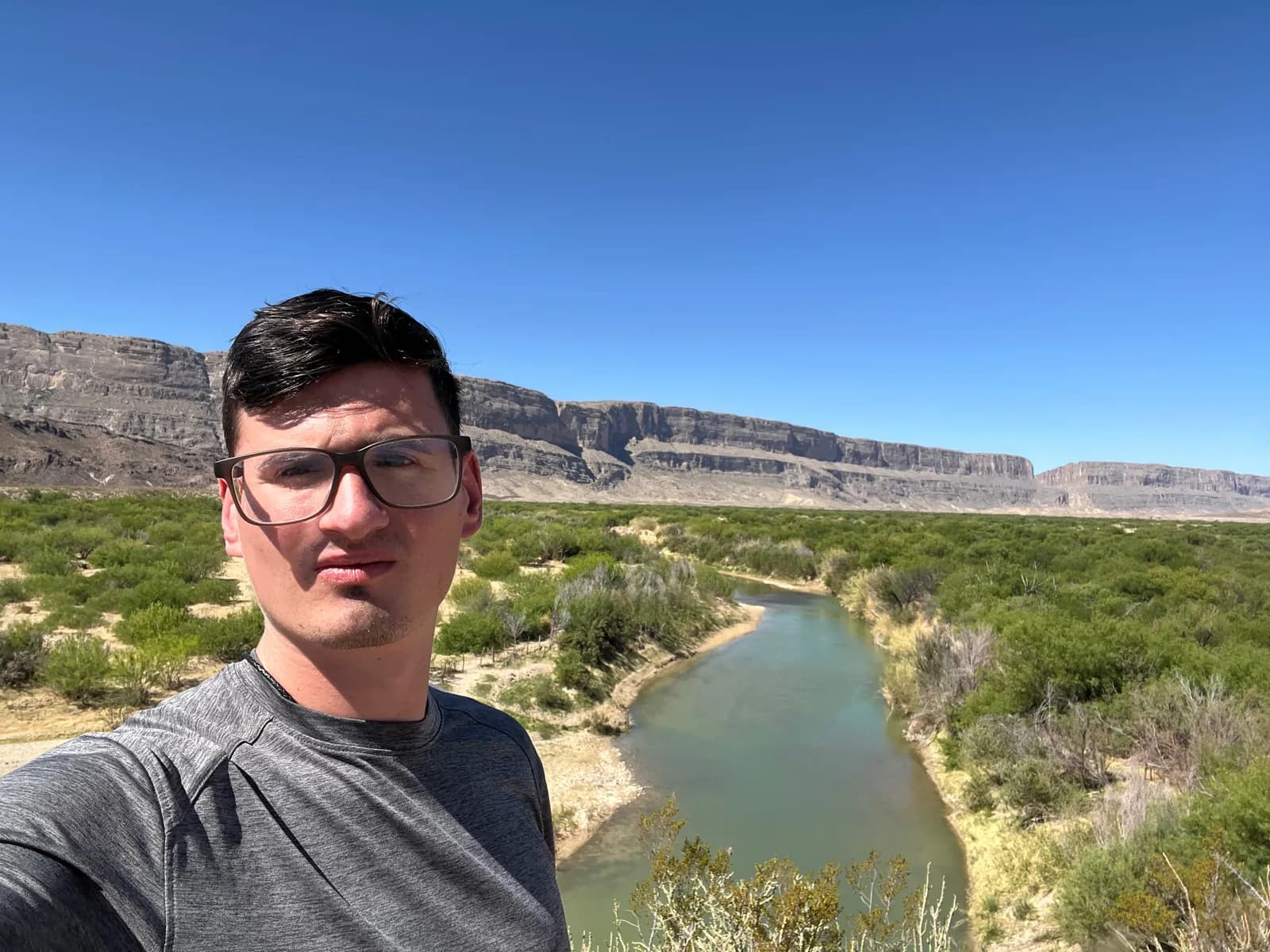 A selfie of a man in glasses and a gray athletic shirt standing in front of a winding river and desert mountains under a blue sky.